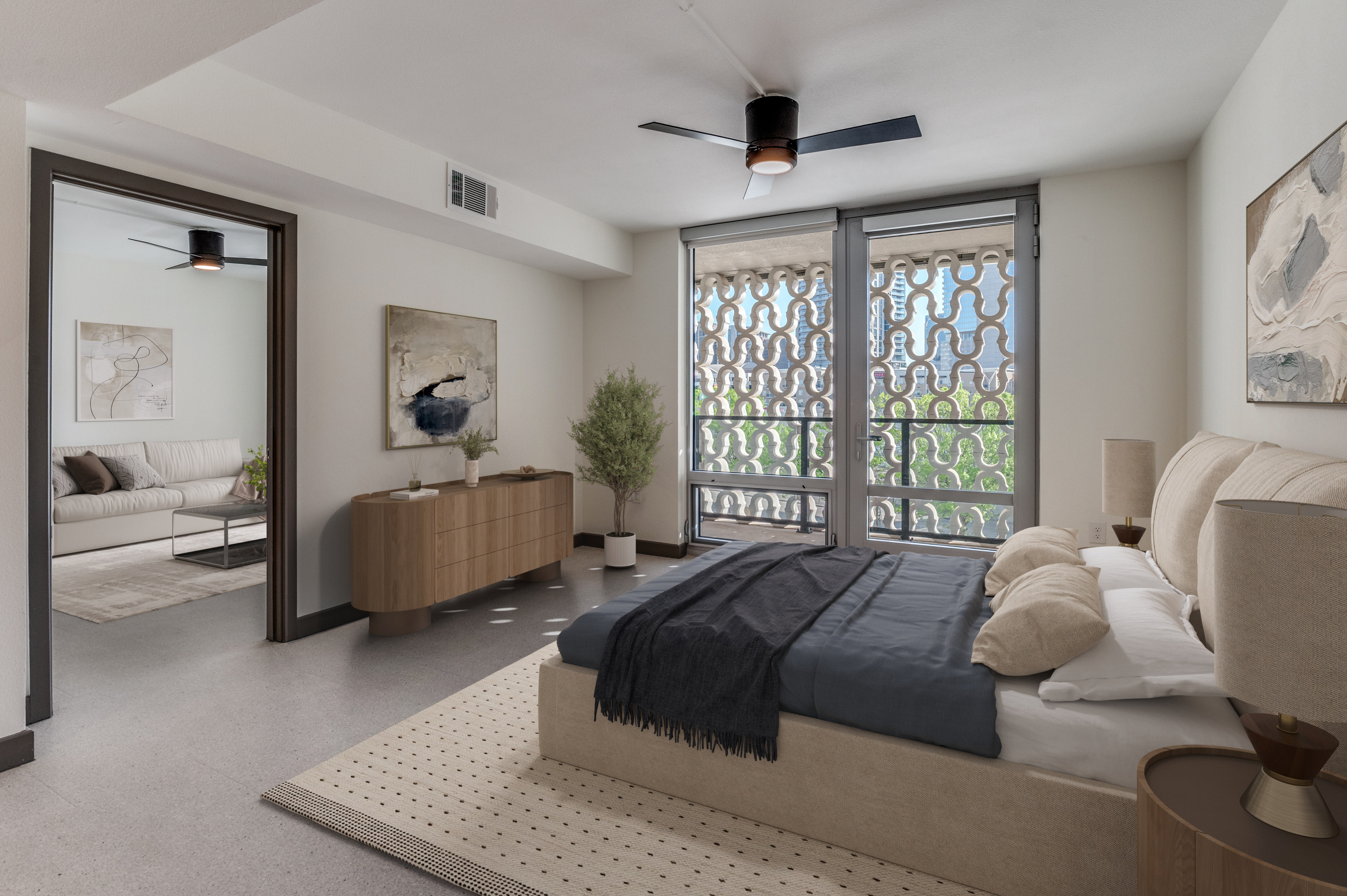 A modern bedroom in one of the finest Dallas Apartments, featuring beige walls, a large bed with a dark blanket, wooden furniture, ceiling fan, artwork, and large windows with patterned shades; an adjoining sitting room is visible.