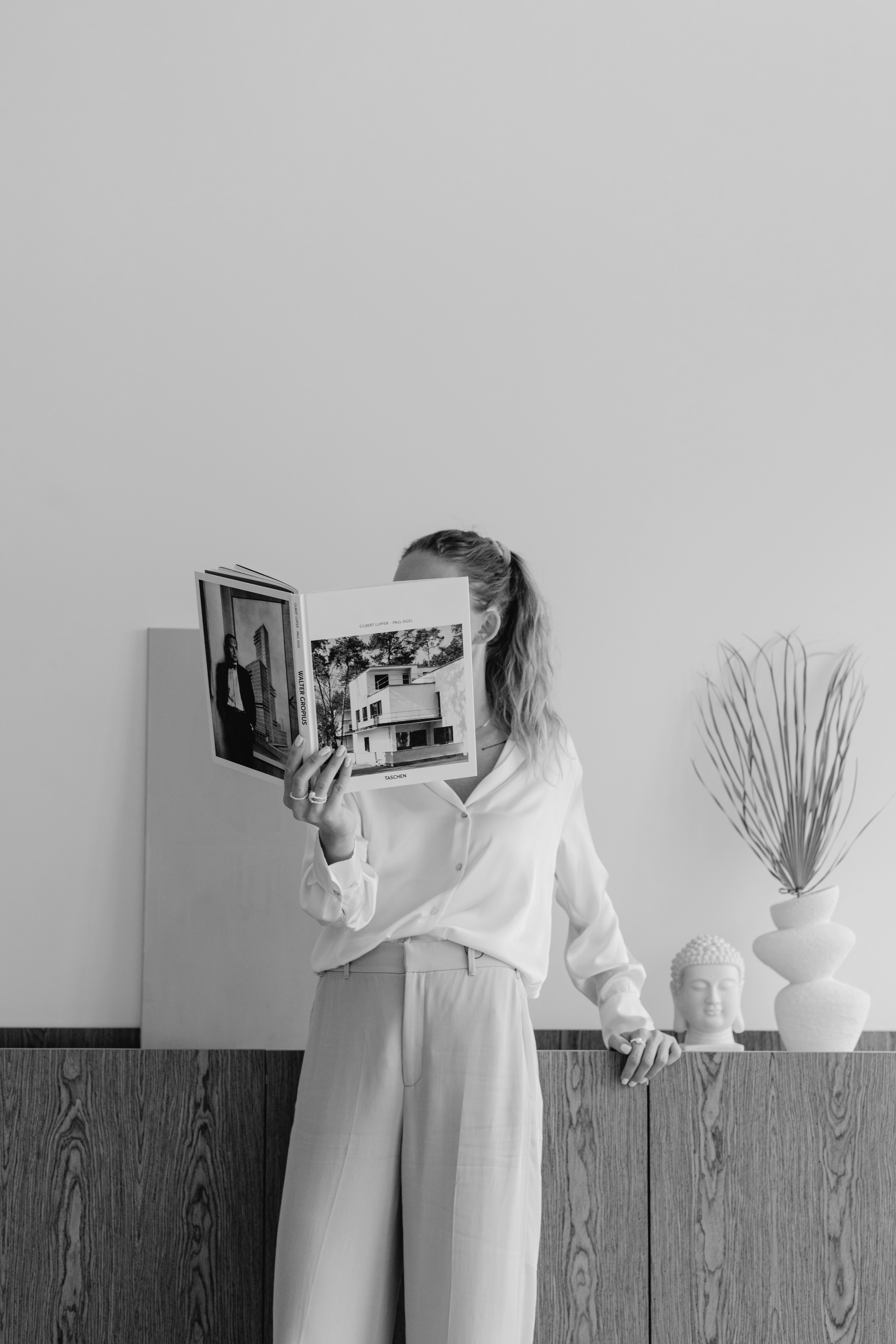 A person in light clothing stands by a wooden cabinet, reading an open magazine; a vase with dry branches and a Buddha head sculpture sit atop the cabinet in this historic Dallas rental.