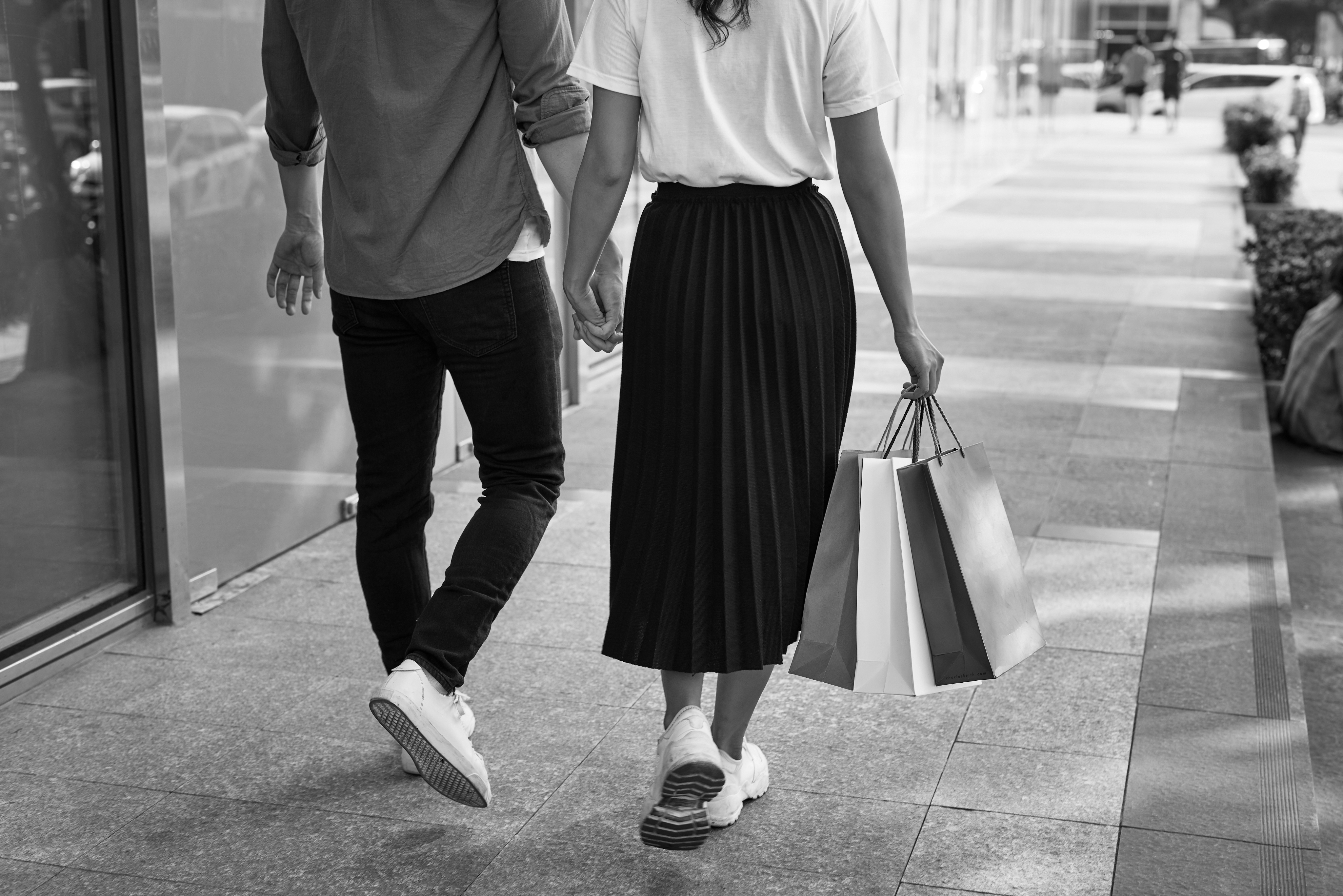 Two people walk hand-in-hand along a sidewalk near Dallas apartments, with one person carrying several shopping bags. The image is in black and white.