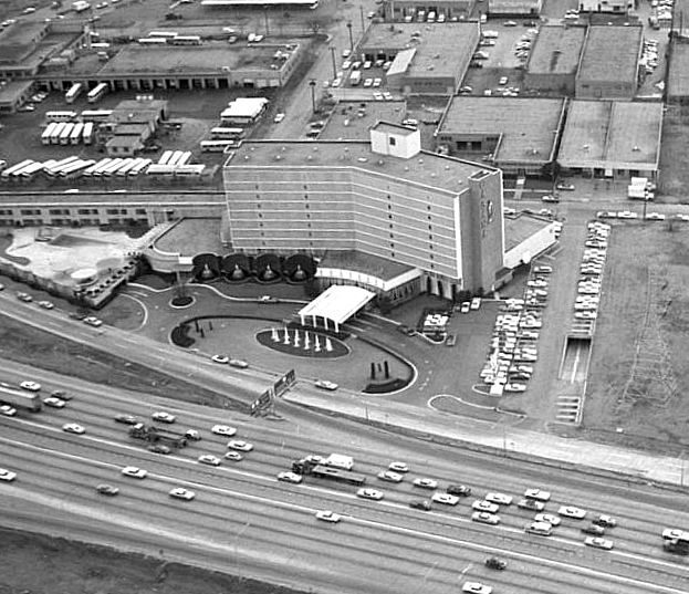 Aerial view of a mid-century hotel with a curved driveway and fountain, surrounded by parking lots, industrial buildings, and Dallas apartments near a busy highway.