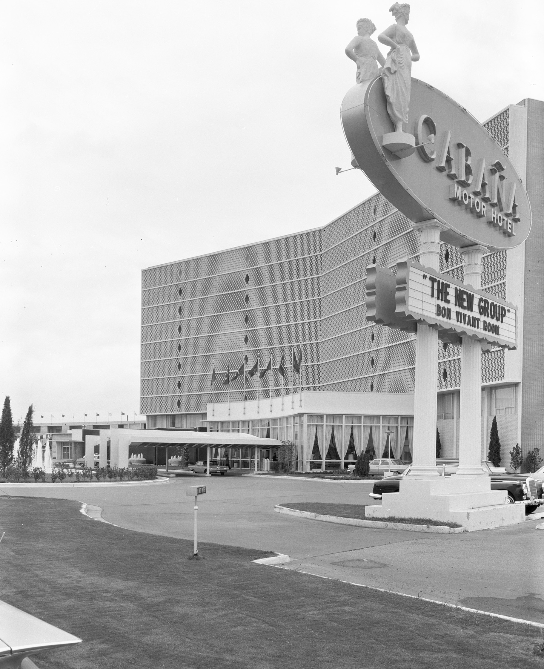 Black and white photo of the historic Cabana Motor Hotel in the Cabana Design District, with a large sign in front advertising "The New Group Bon Vivant Room" and a multi-story building in the background.