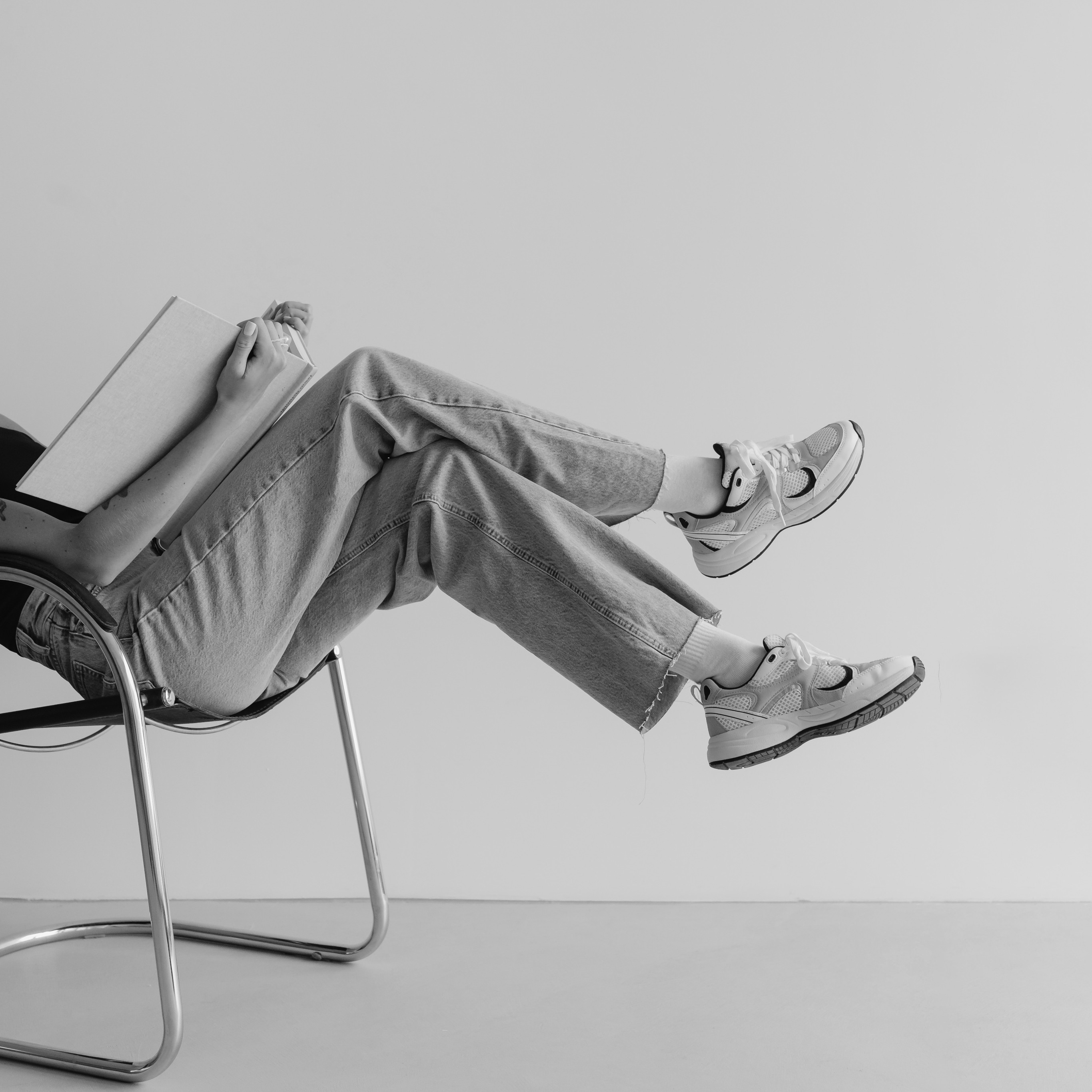 Person sitting on a chair with legs crossed, holding an open book. They wear jeans, socks, and sneakers. The plain, minimalistic background echoes the modern vibe of Cabana Design District apartments in Dallas.