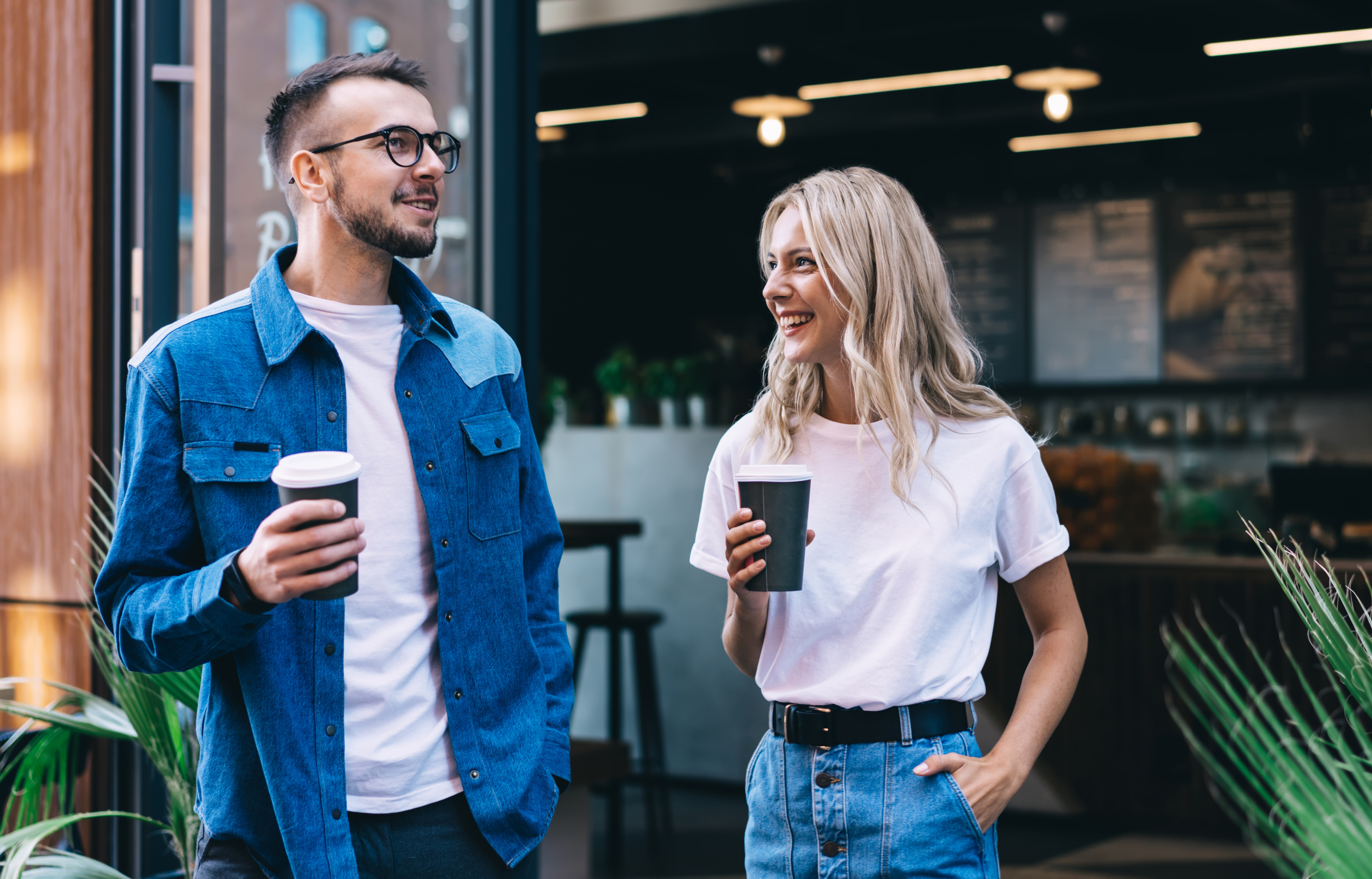 A man and a woman holding coffee cups stand outside a café in Dallas, smiling and talking. Both wear casual outfits with blue jeans and white shirts—typical of friends enjoying city life near their modern Dallas Apartments.