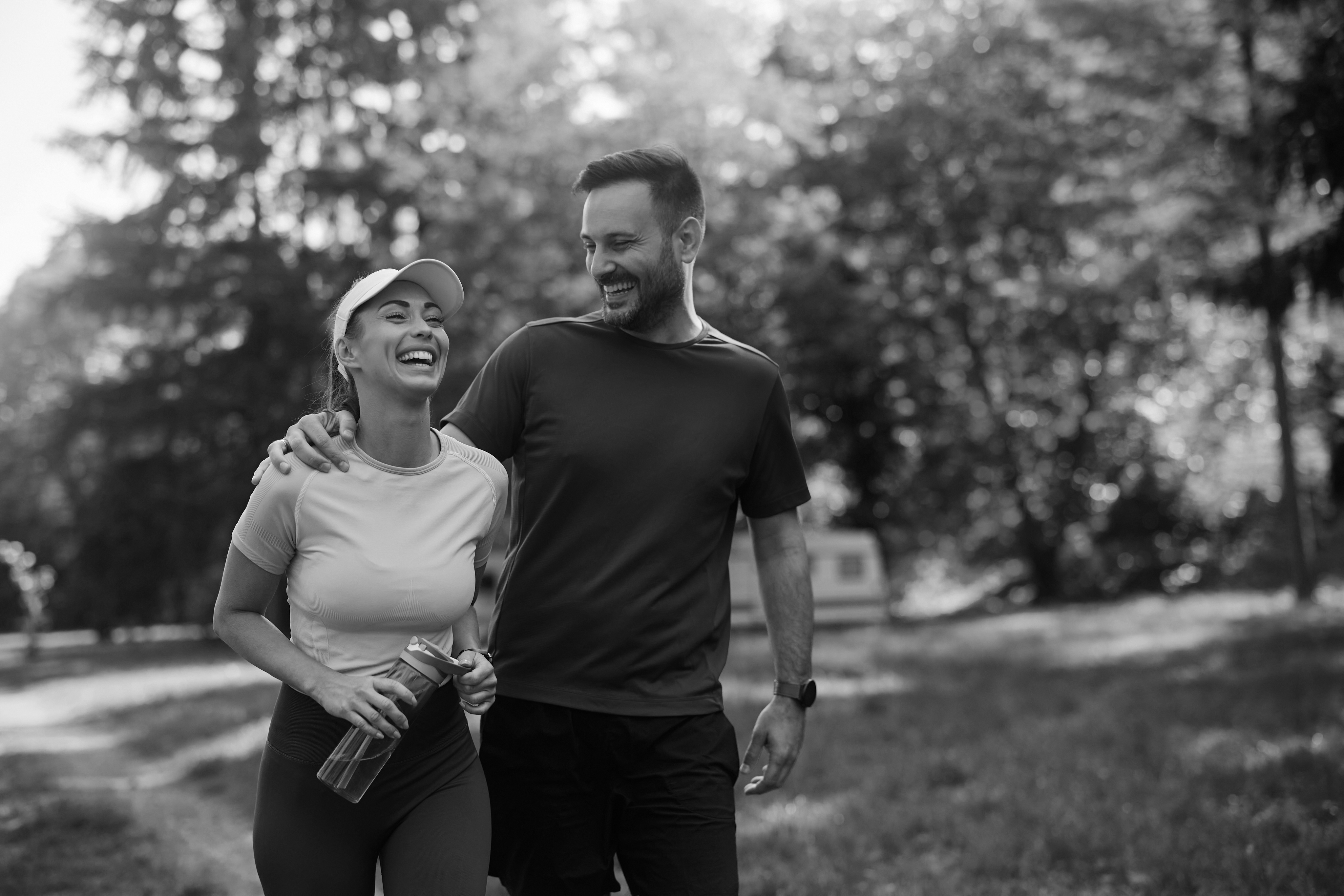 A woman and man walk outdoors on a sunny Dallas day, smiling and laughing together. The woman holds a water bottle and wears a cap. Trees and grass are visible in the background near a historic Dallas rental.