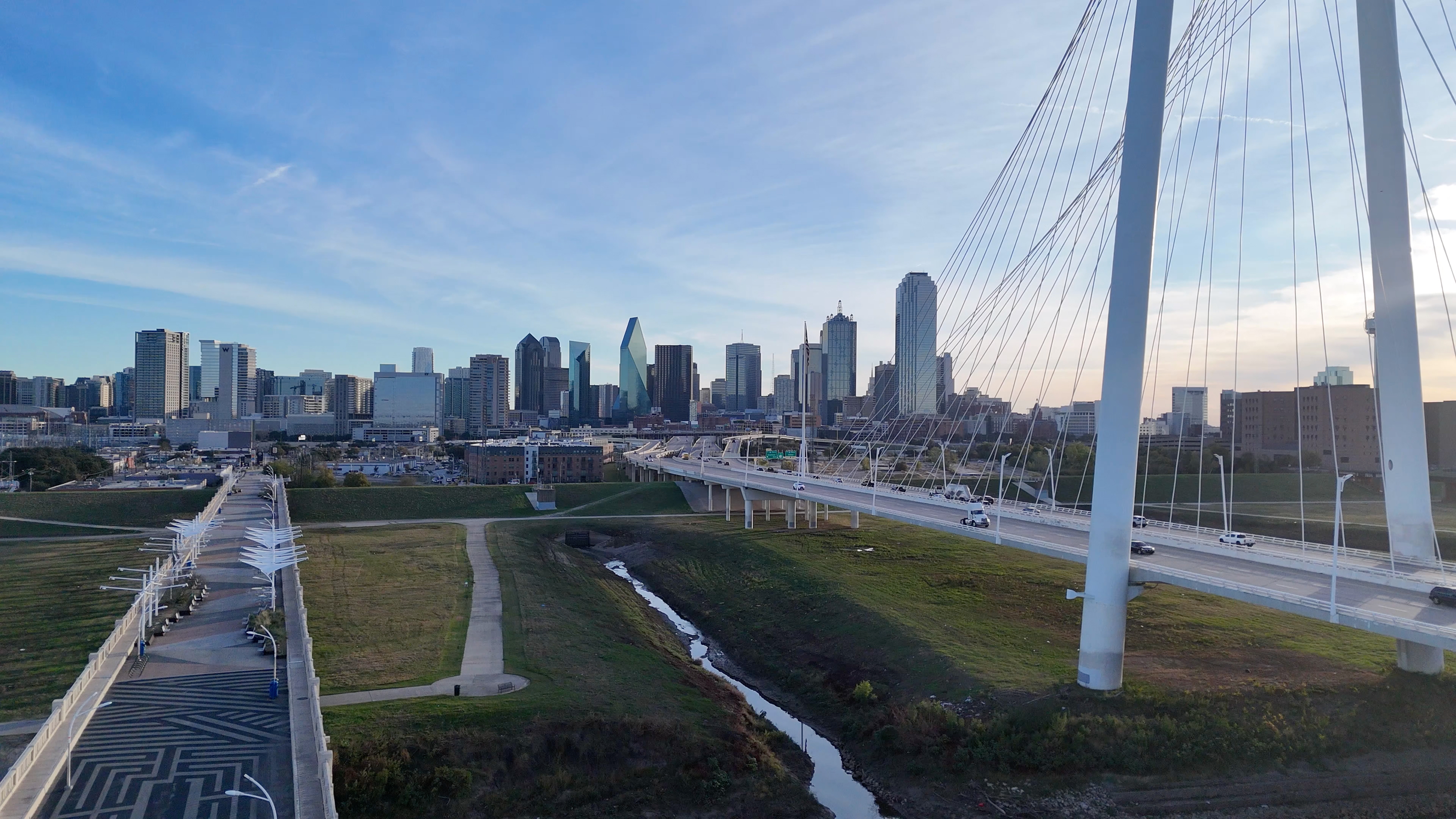 A cable-stayed bridge with white cables spans a grassy area toward a city skyline of modern high-rise buildings under a blue sky, reflecting the vibrant ambiance of the nearby Cabana Design District.