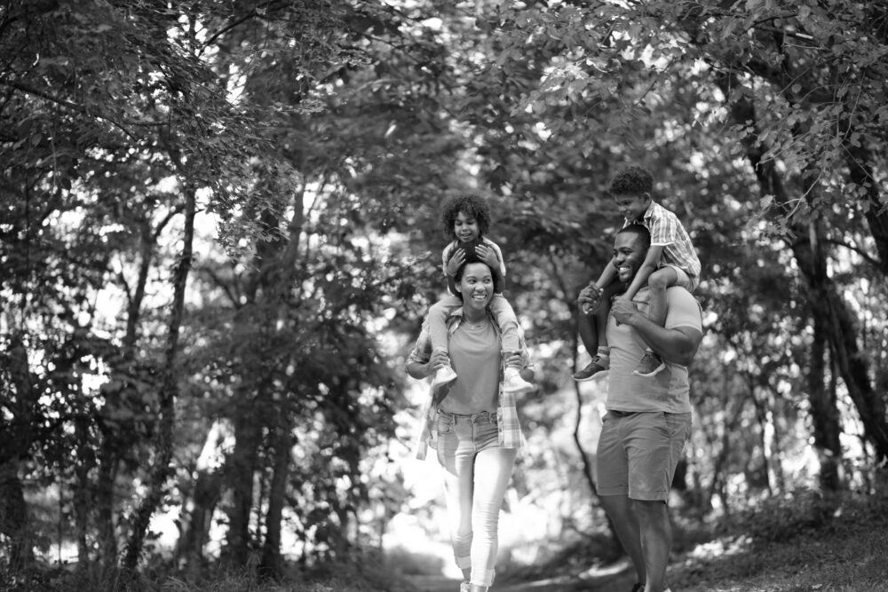 A family of four enjoys a walk outdoors near Dallas Apartments, with two adults carrying children on their shoulders, surrounded by trees.