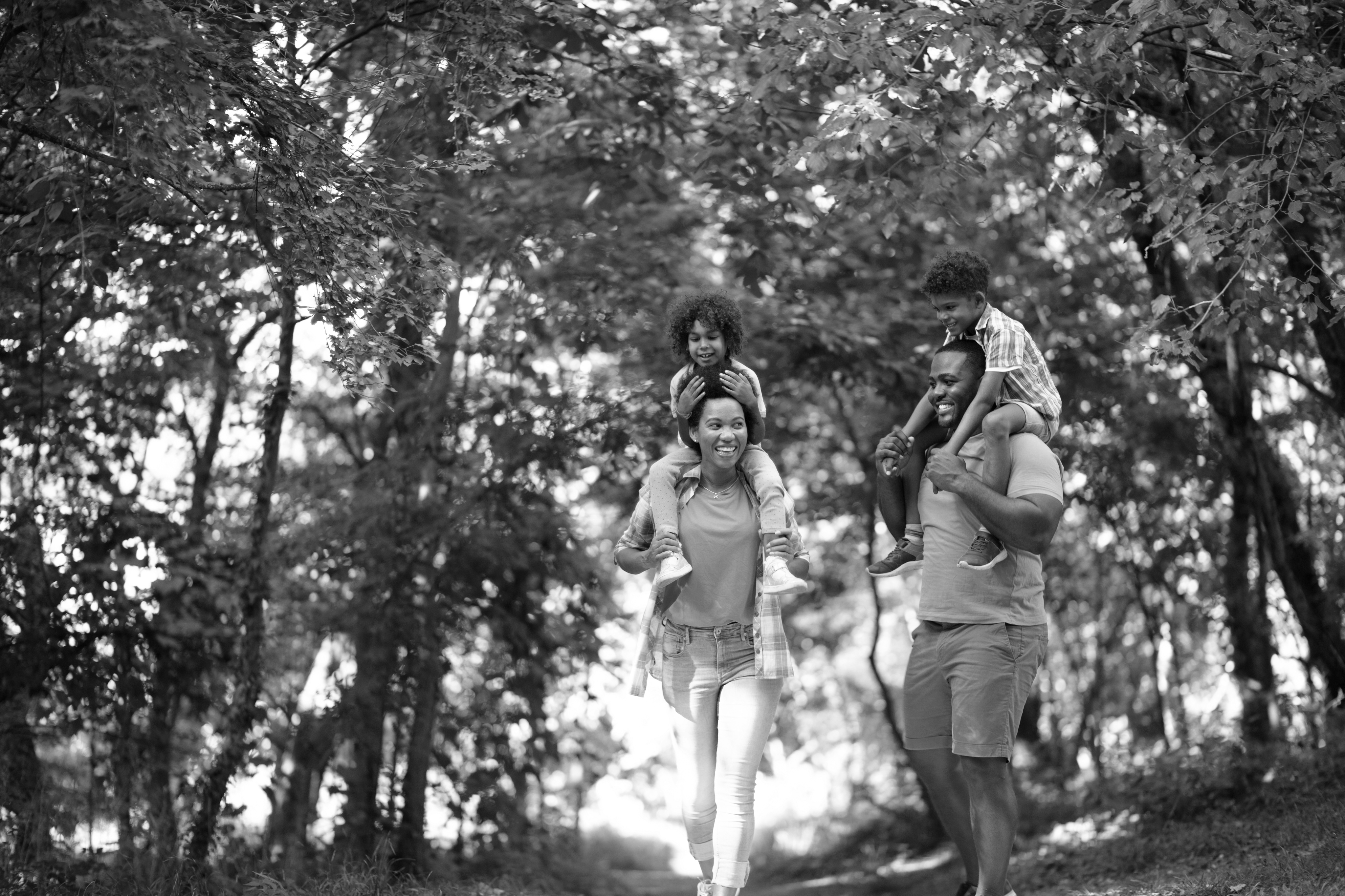 A family of four enjoys a walk outdoors near Dallas Apartments, with two adults carrying children on their shoulders, surrounded by trees.