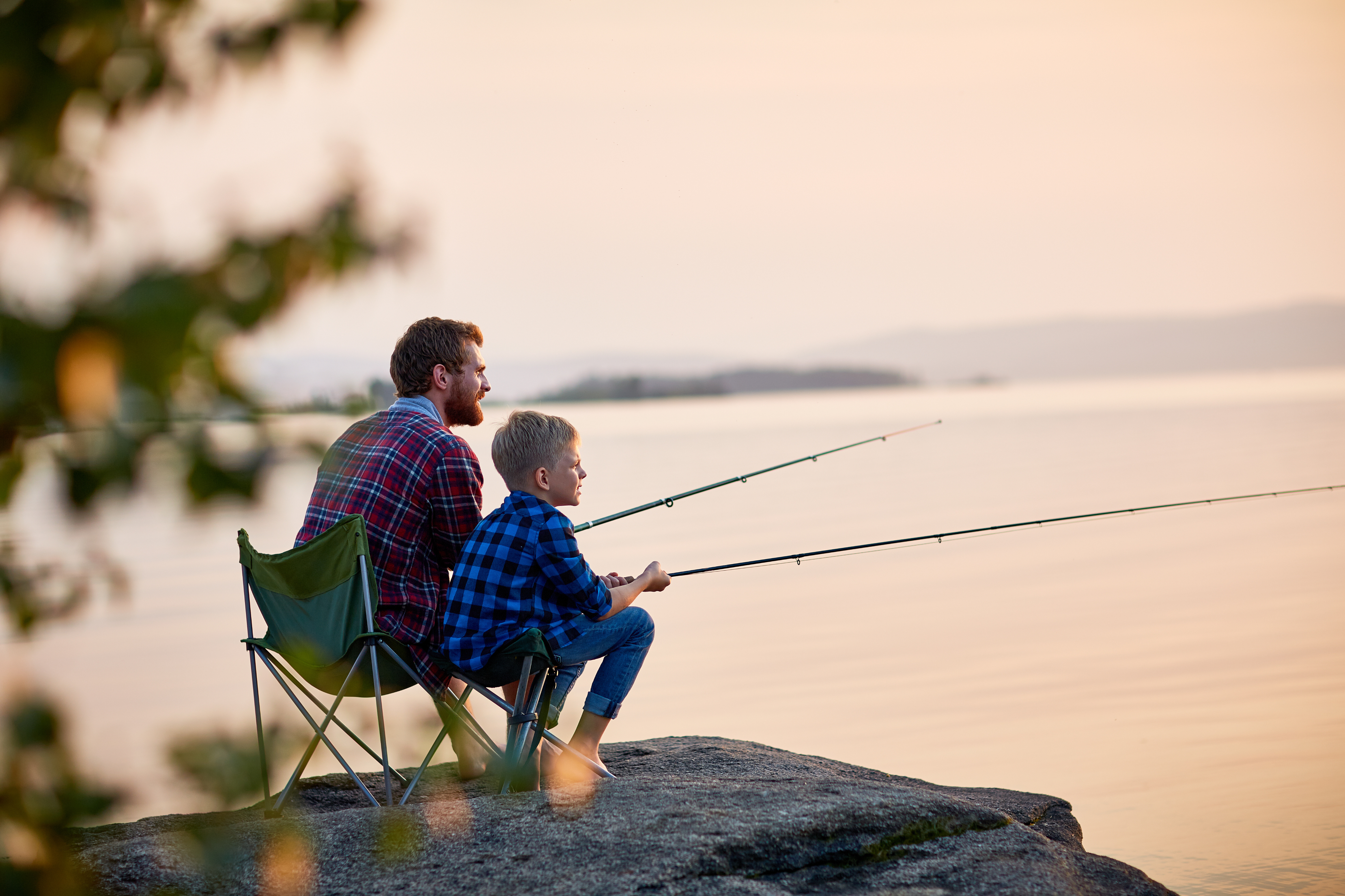 An adult and a child sit on a rock by a lake, fishing with rods at sunset. Both wear plaid shirts and look towards the water, enjoying peaceful moments away from their Dallas Apartments.