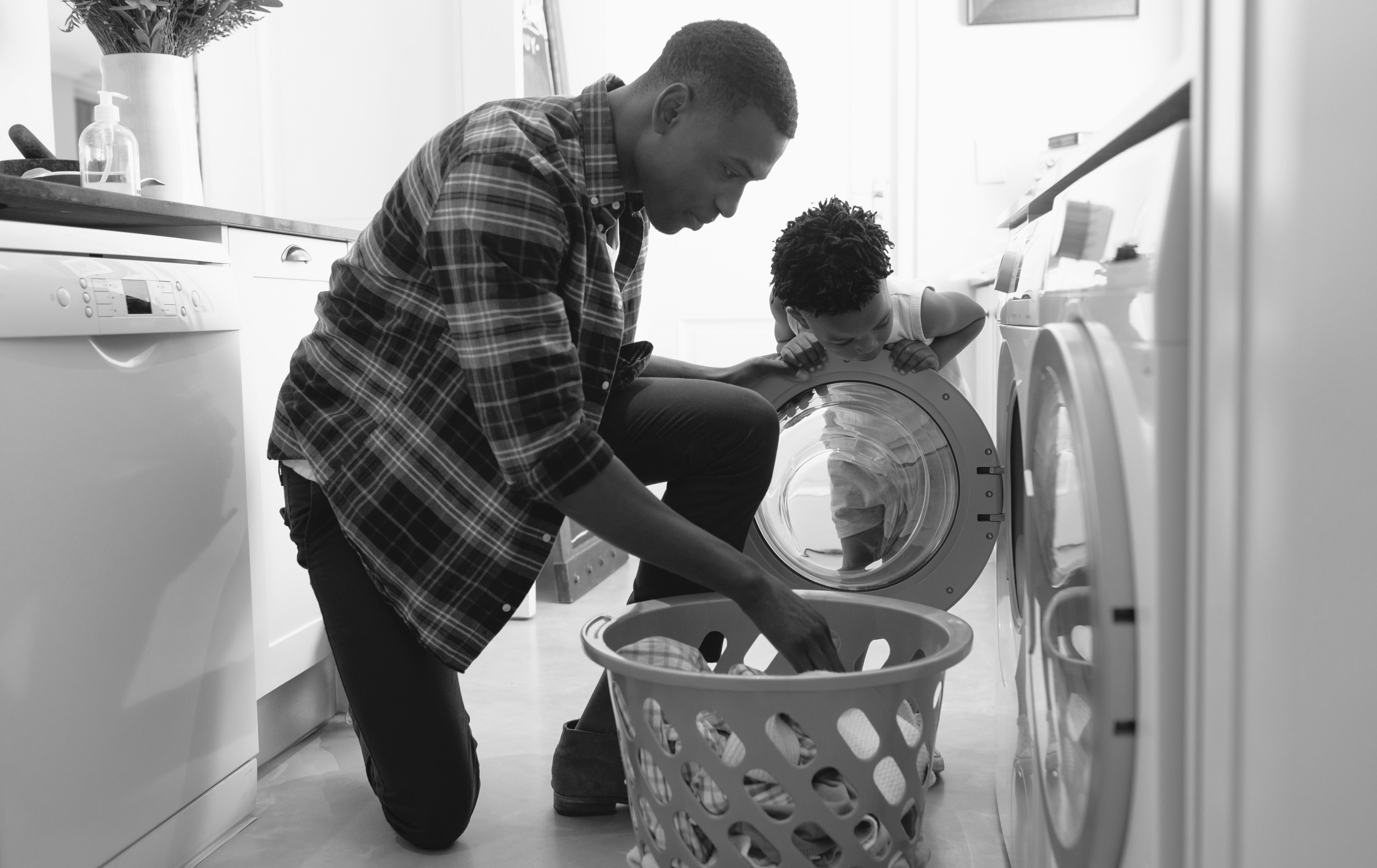 In a stylish Cabana Design District apartment, a man kneels by the washing machine, placing clothes inside as a young child curiously peers into the open drum.