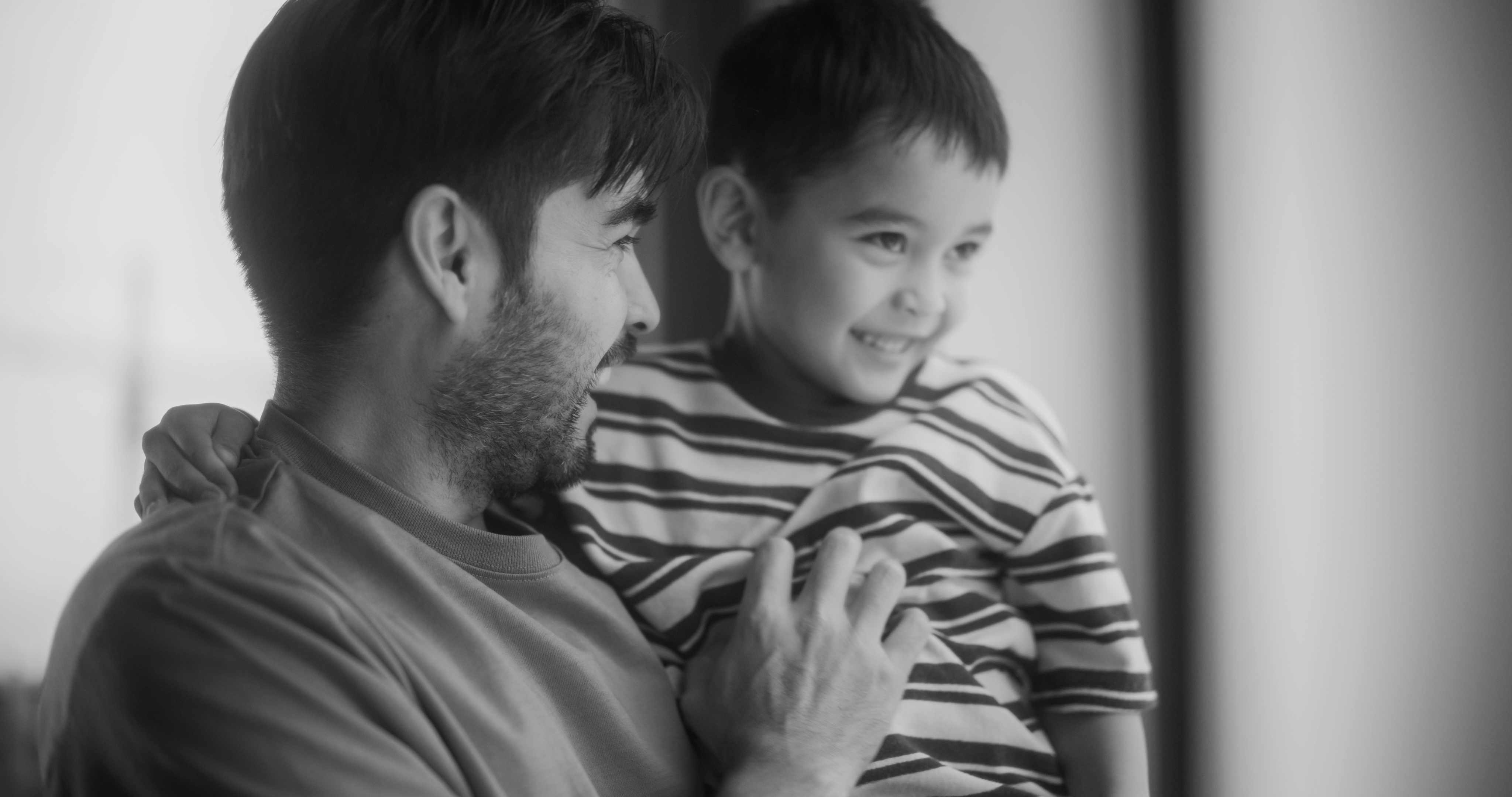 A man holds a smiling young boy in his arms as they face each other in a brightly lit indoor setting, capturing a warm moment in the heart of Dallas’s Historic Cabana Design District.