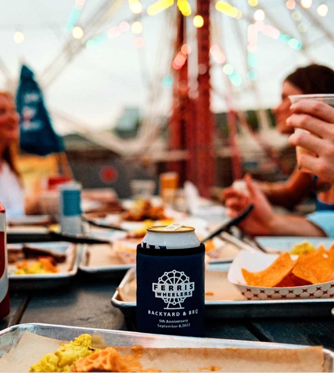A group of people eat at an outdoor table with food trays and drinks near Cabana Design District Apartments in Dallas; a Ferris Wheel and string lights are visible in the background.