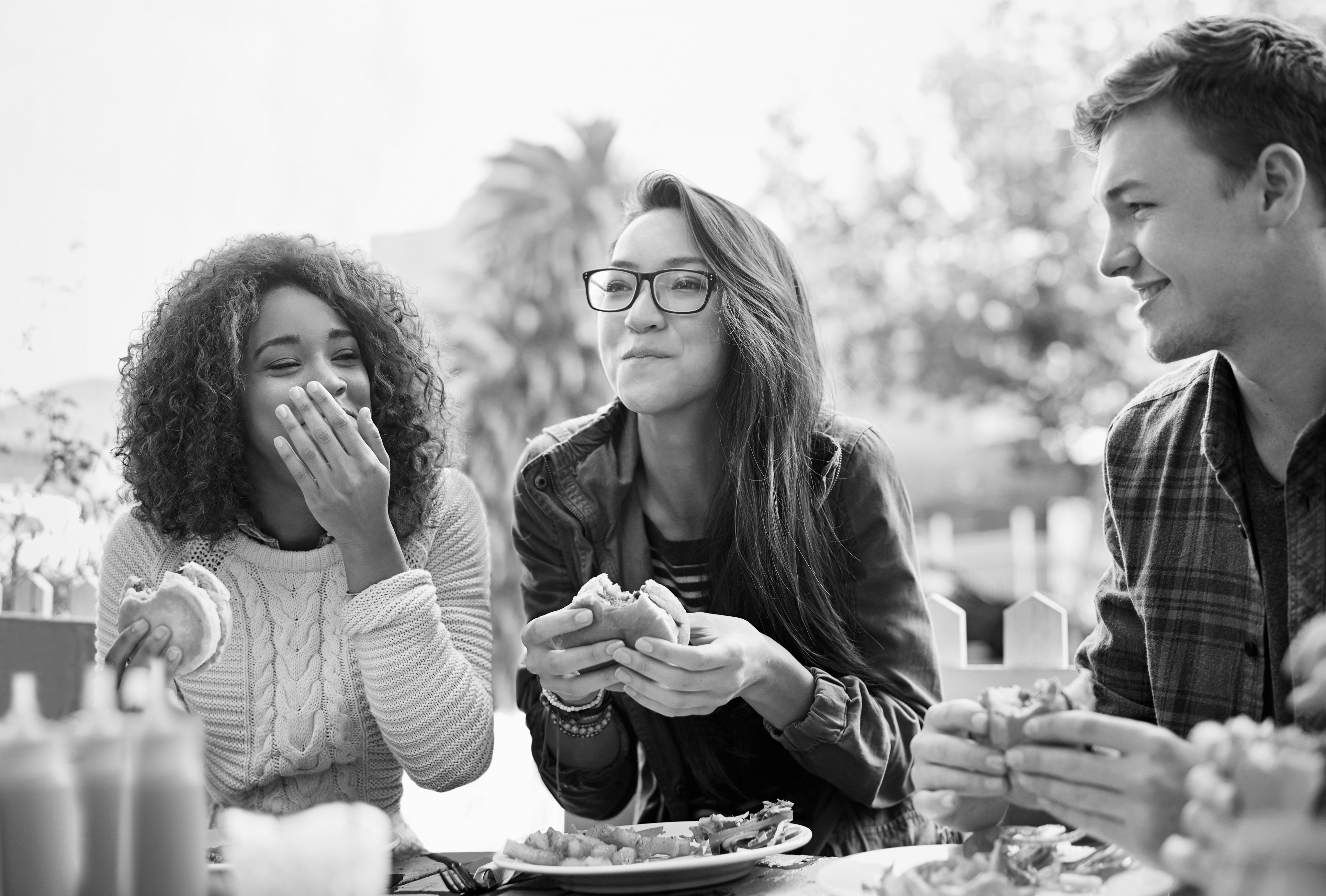 Three people sit at an outdoor table, eating sandwiches and smiling, with bottles of condiments visible in the foreground at Cabana Design District Apartments in Dallas.