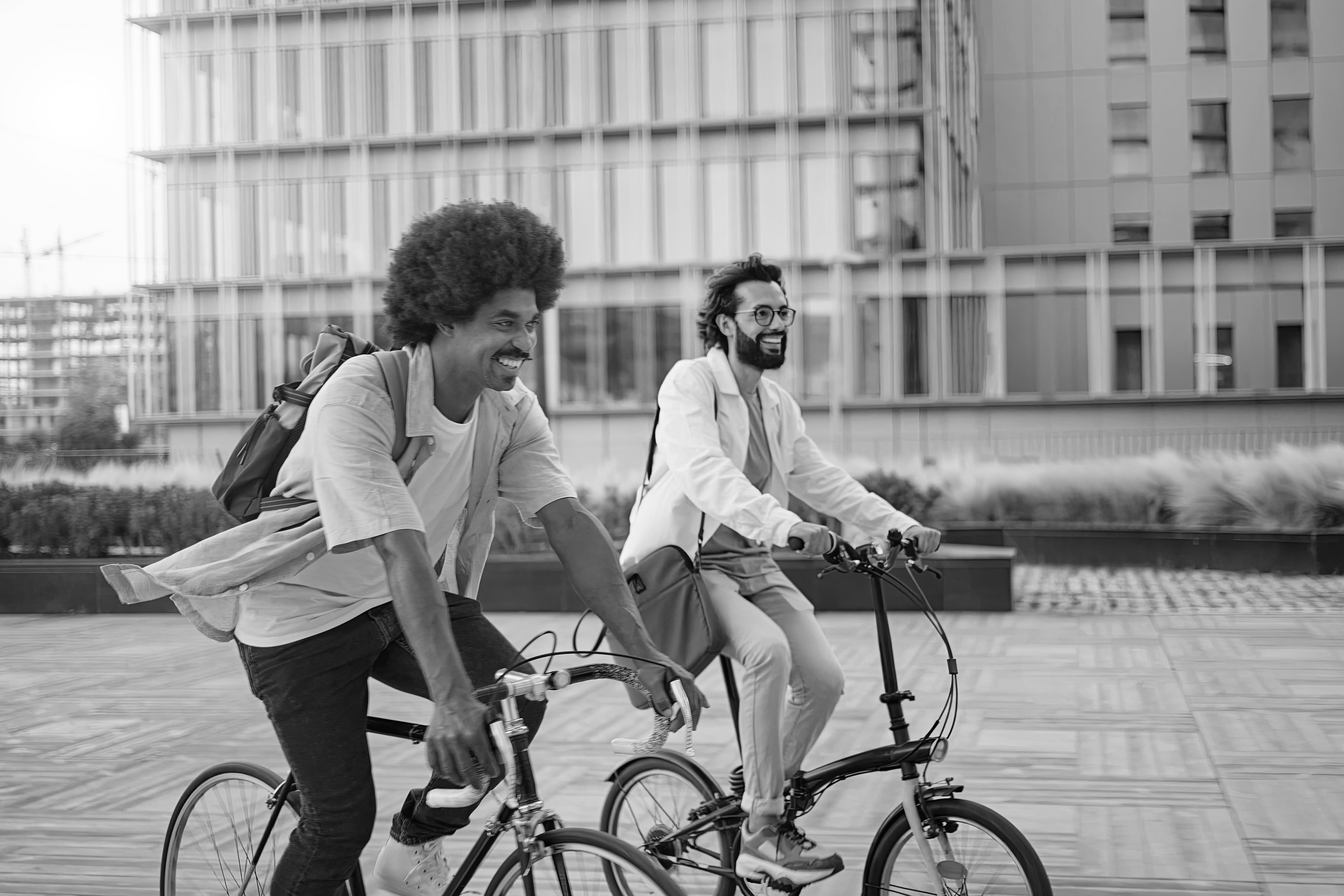 Two men riding bicycles side by side on a paved path in an urban area, both smiling and carrying backpacks, with historic Dallas Apartments visible in the background.