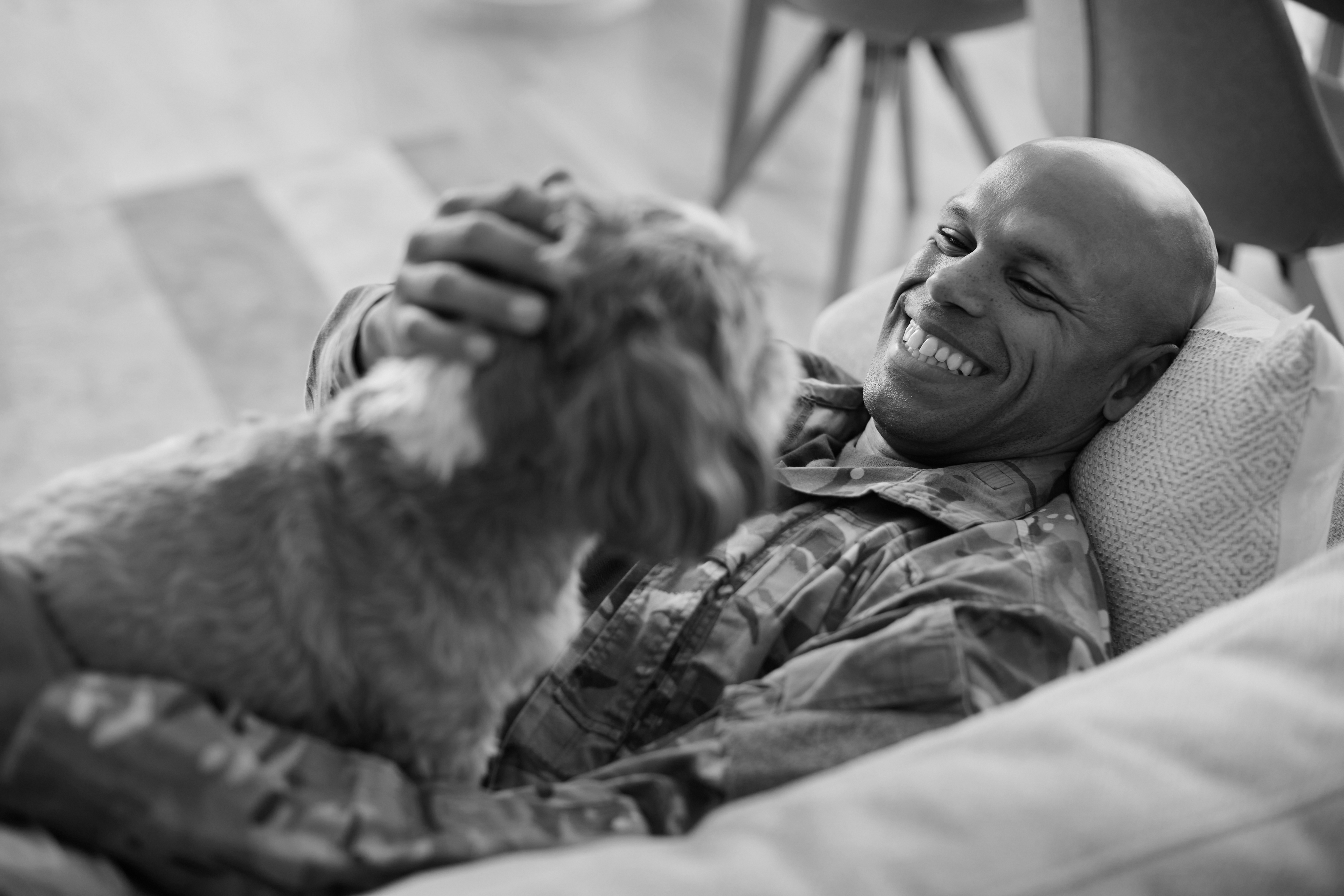 A man in military uniform lies on a couch inside a historic Dallas apartment, smiling and petting a small dog that is sitting on his chest.