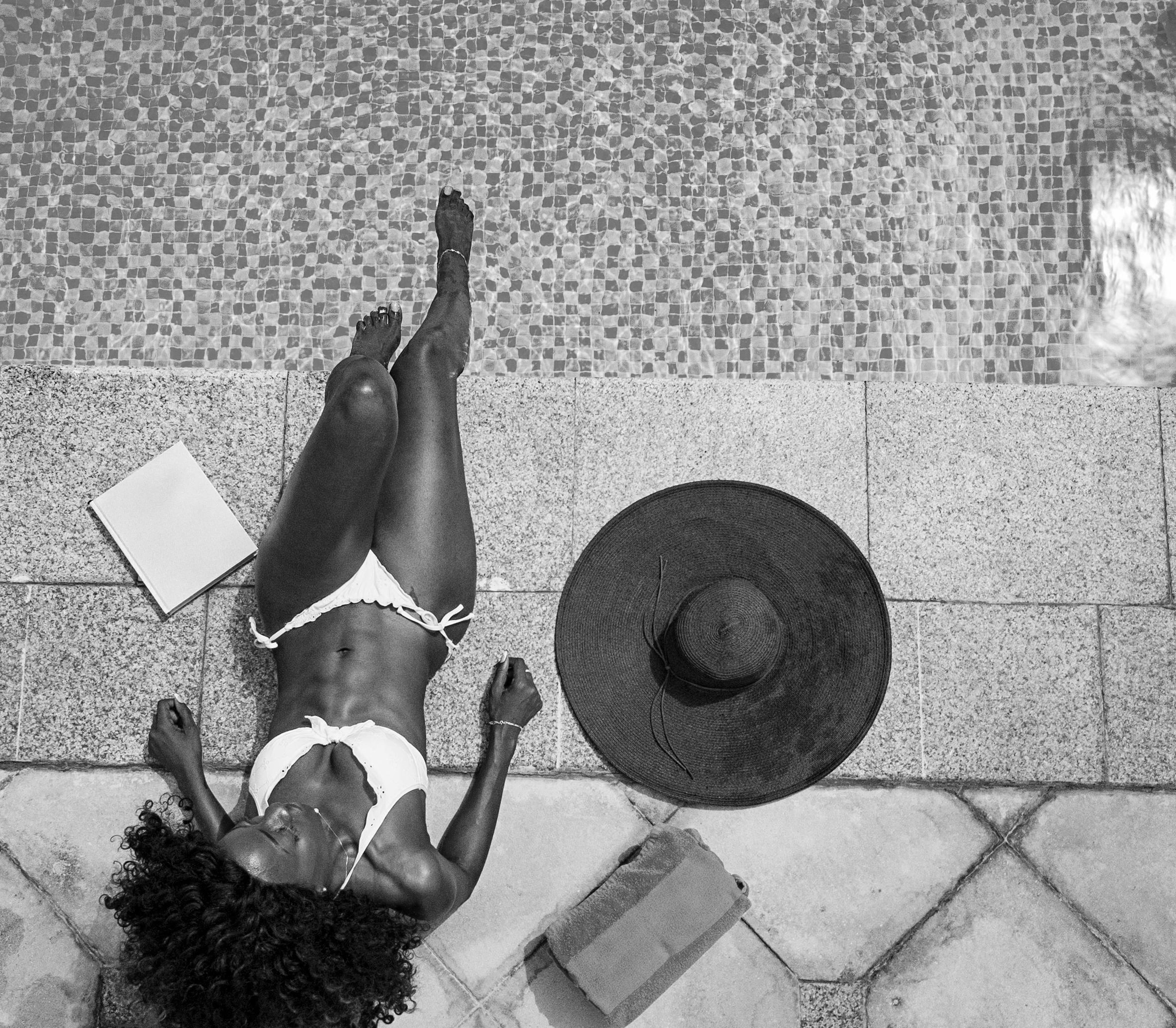 A woman in a white bikini relaxes on a poolside deck at a Dallas Rental, with a book, black wide-brimmed hat, and towel beside her, near the edge of the tiled Cabana Design District swimming pool.