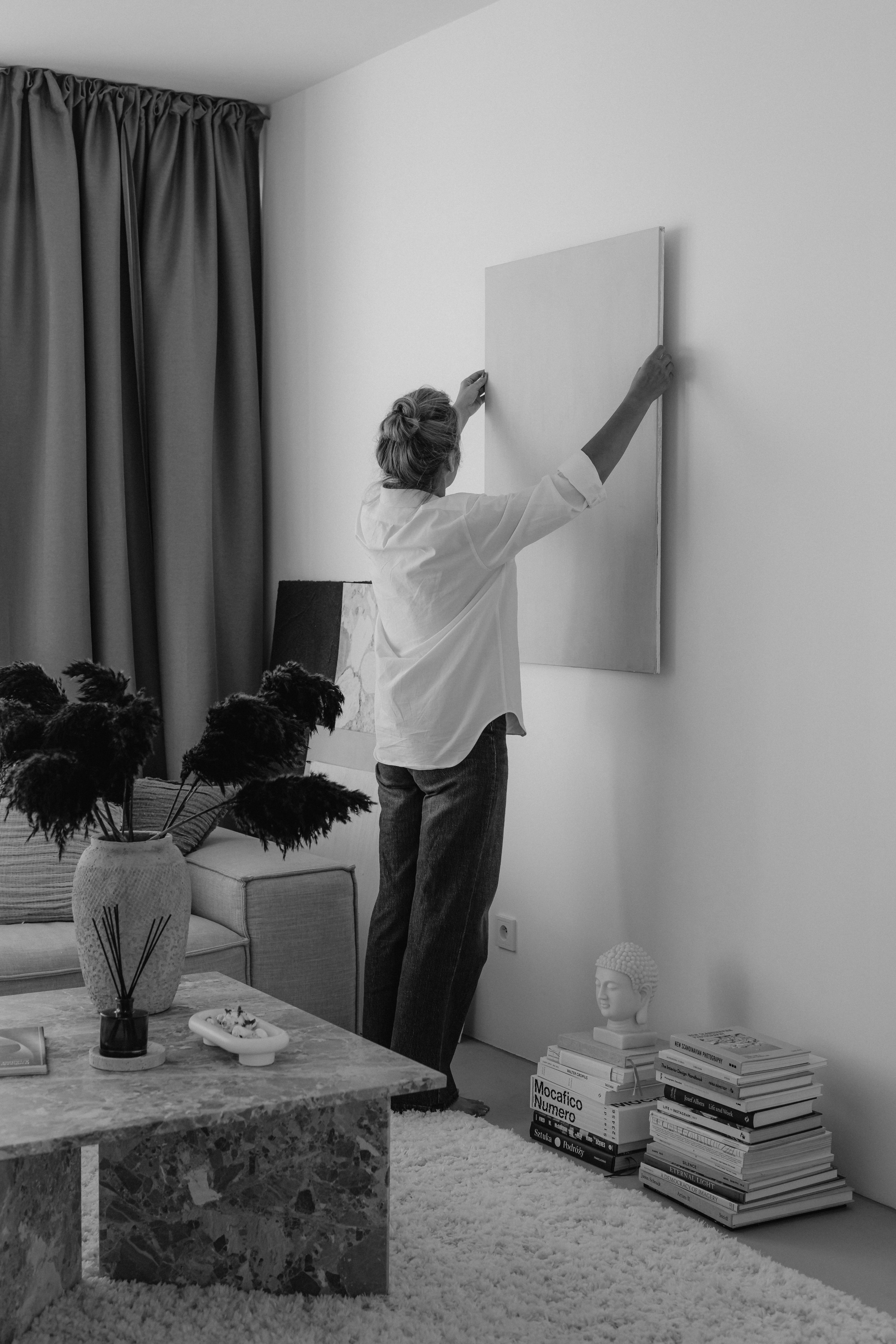A person hangs a blank canvas on the wall of a living room in the historic Cabana Design District, featuring a marble coffee table, books, a small sculpture, and a vase with dark foliage.