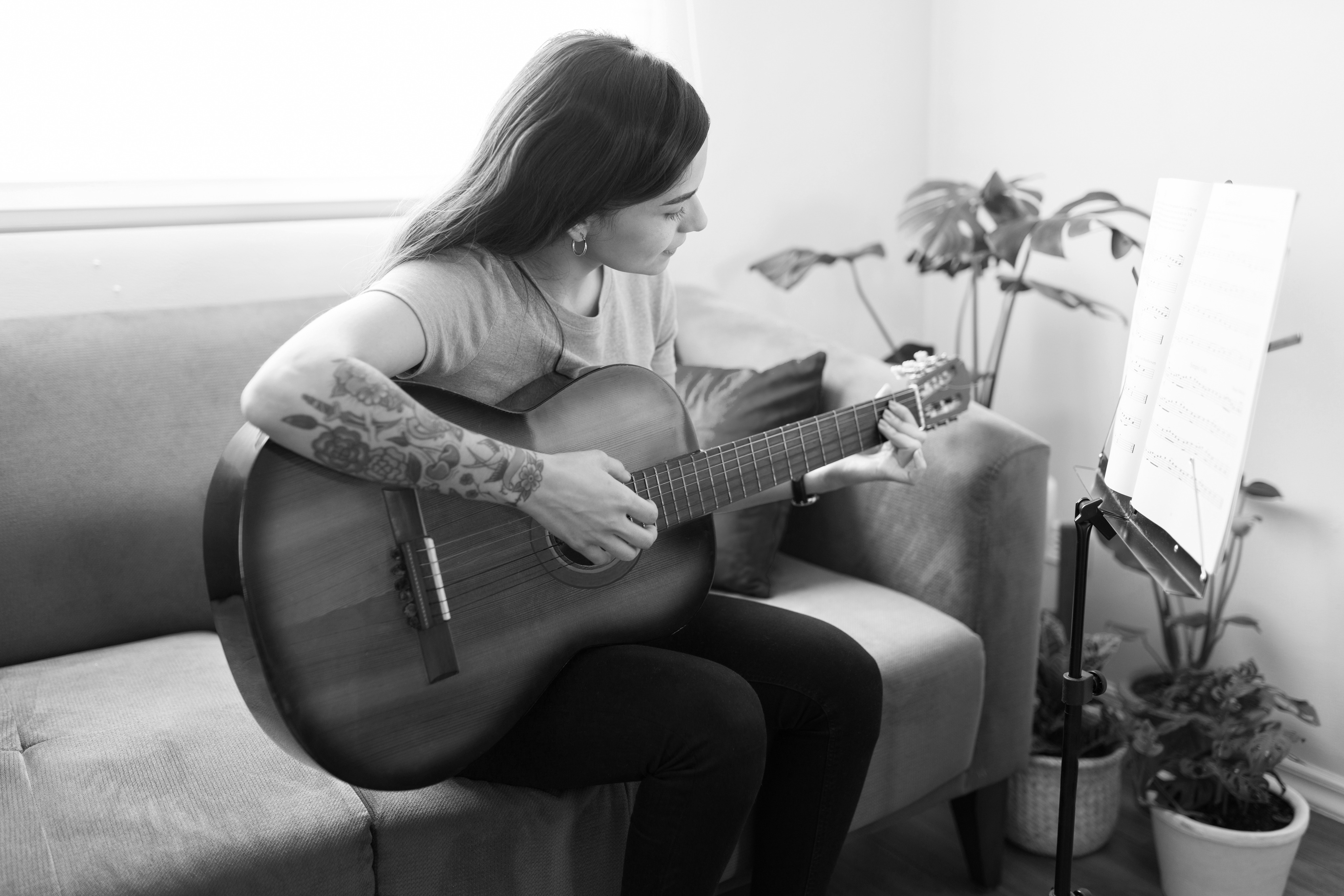 A person with tattoos sits on a couch in their Dallas apartment, playing an acoustic guitar and reading sheet music from a stand, surrounded by houseplants—perfect for unwinding at Cabana Design District.