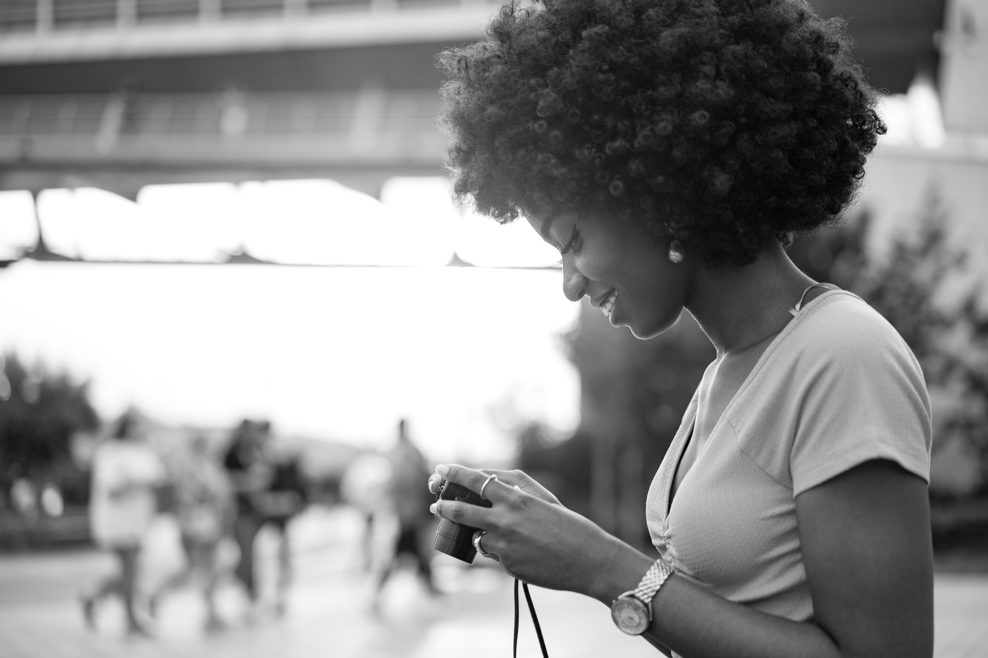 A woman stands outdoors in Dallas, looking down and smiling while holding a camera. People walk in the blurred background beneath a bridge. The black-and-white image captures the charm of city life near popular Dallas apartments.