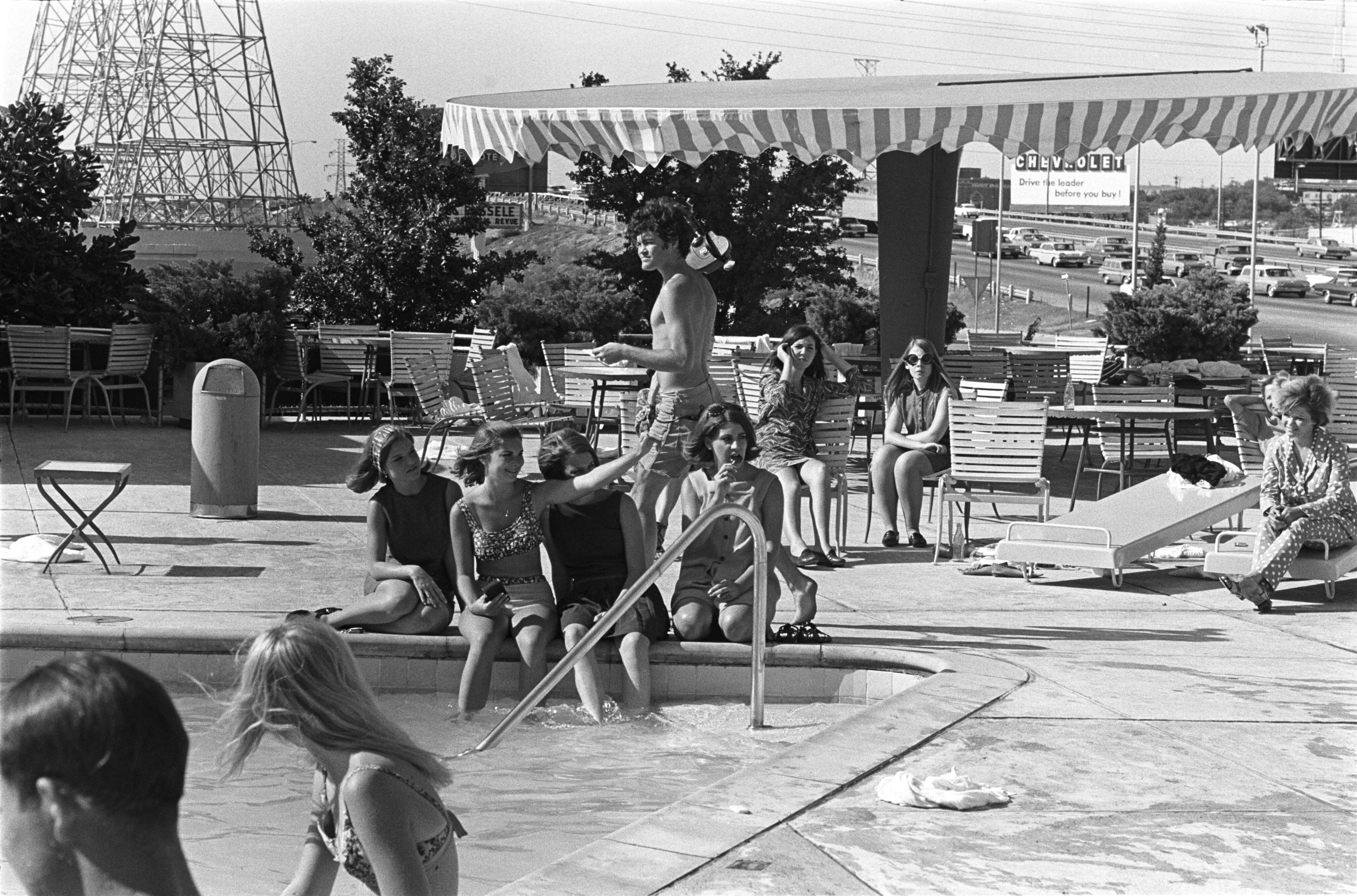 A group of people sit by a pool at Historic Apartments, some with their feet in the water, while others relax on lounge chairs under a striped canopy near the Cabana Design District’s busy road.