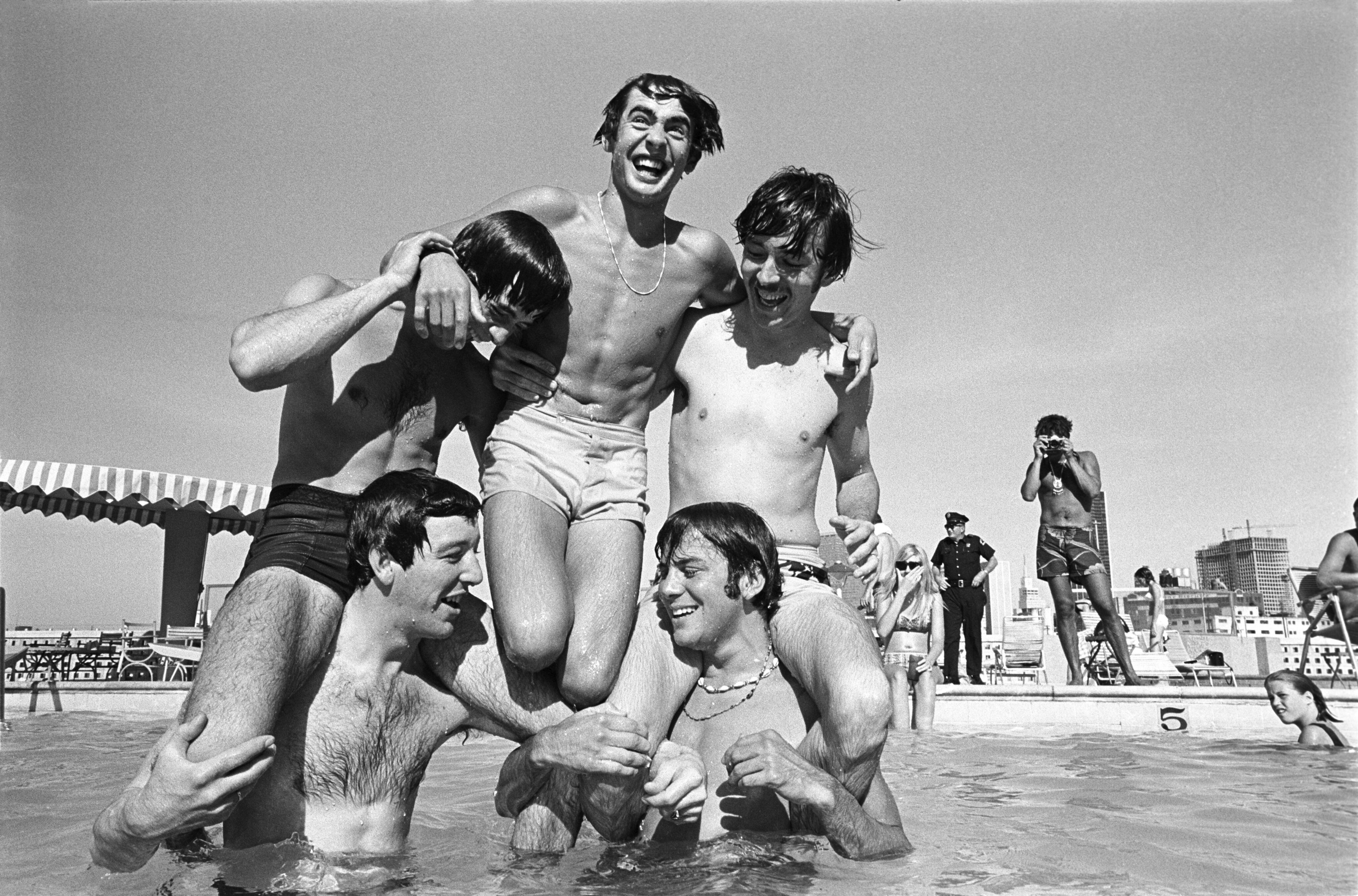 Five young men in swim trunks laugh in a swimming pool at Cabana Design District, some sitting on others' shoulders, with people and city buildings—hallmarks of Dallas Apartments—visible in the background.