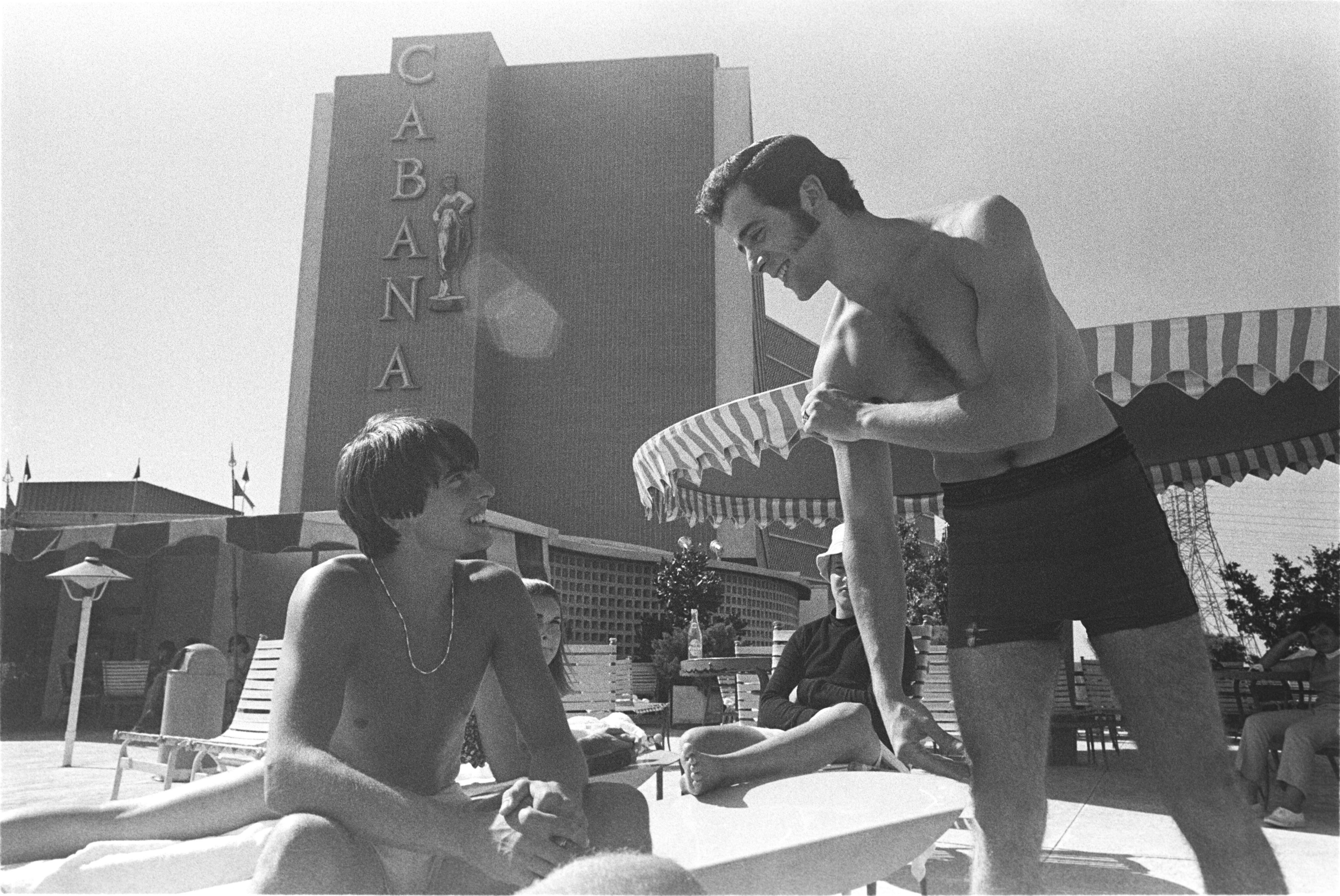 Two men in swim trunks interact by a poolside at the Cabana Design District, with the hotel building and sunbathers visible in the background on a sunny day—an ideal scene for those seeking vibrant Dallas apartments.