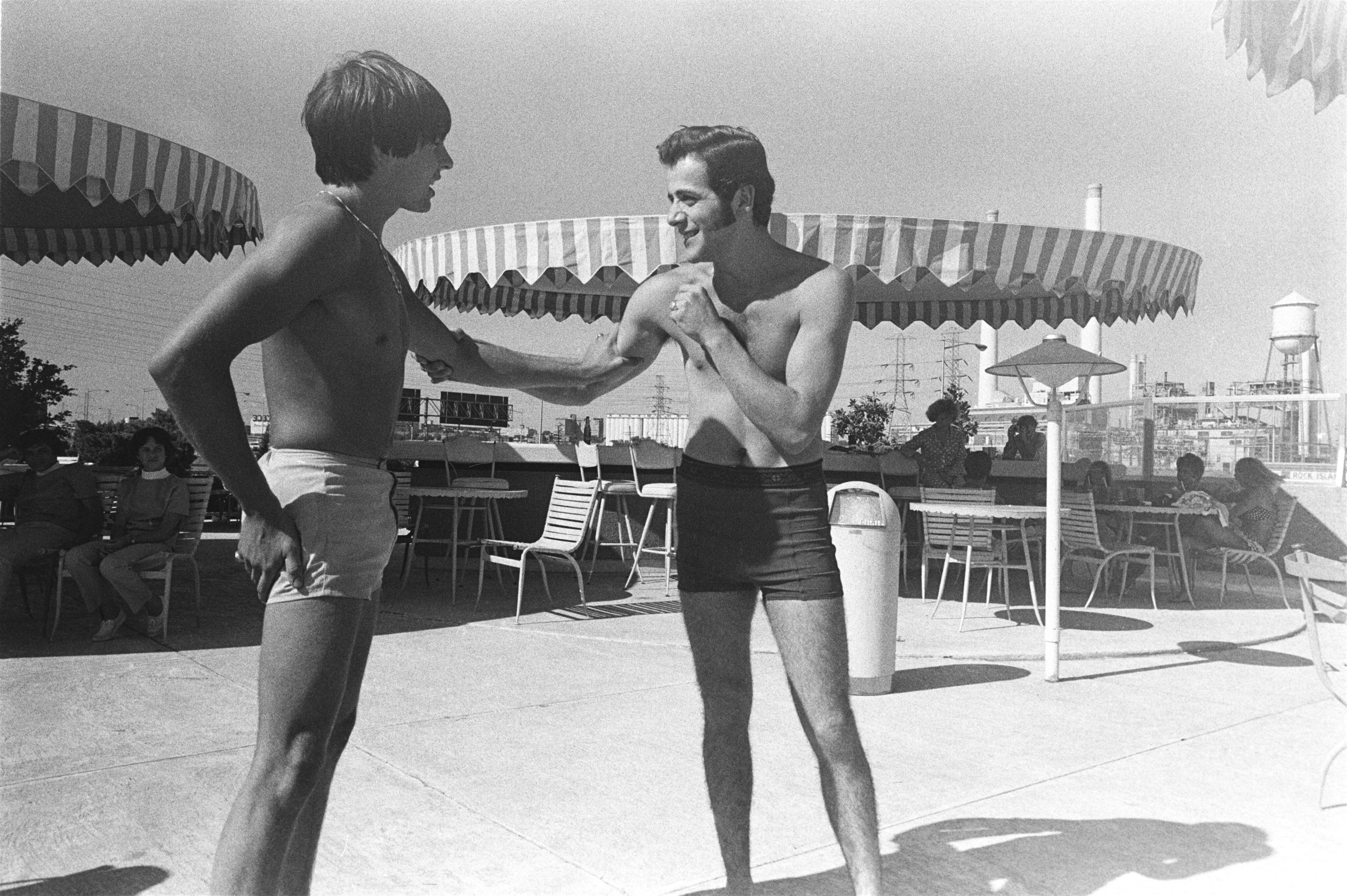 Two men in swim trunks stand and interact by poolside lounge chairs and umbrellas at Dallas apartments, with people relaxing in the background on a sunny day.