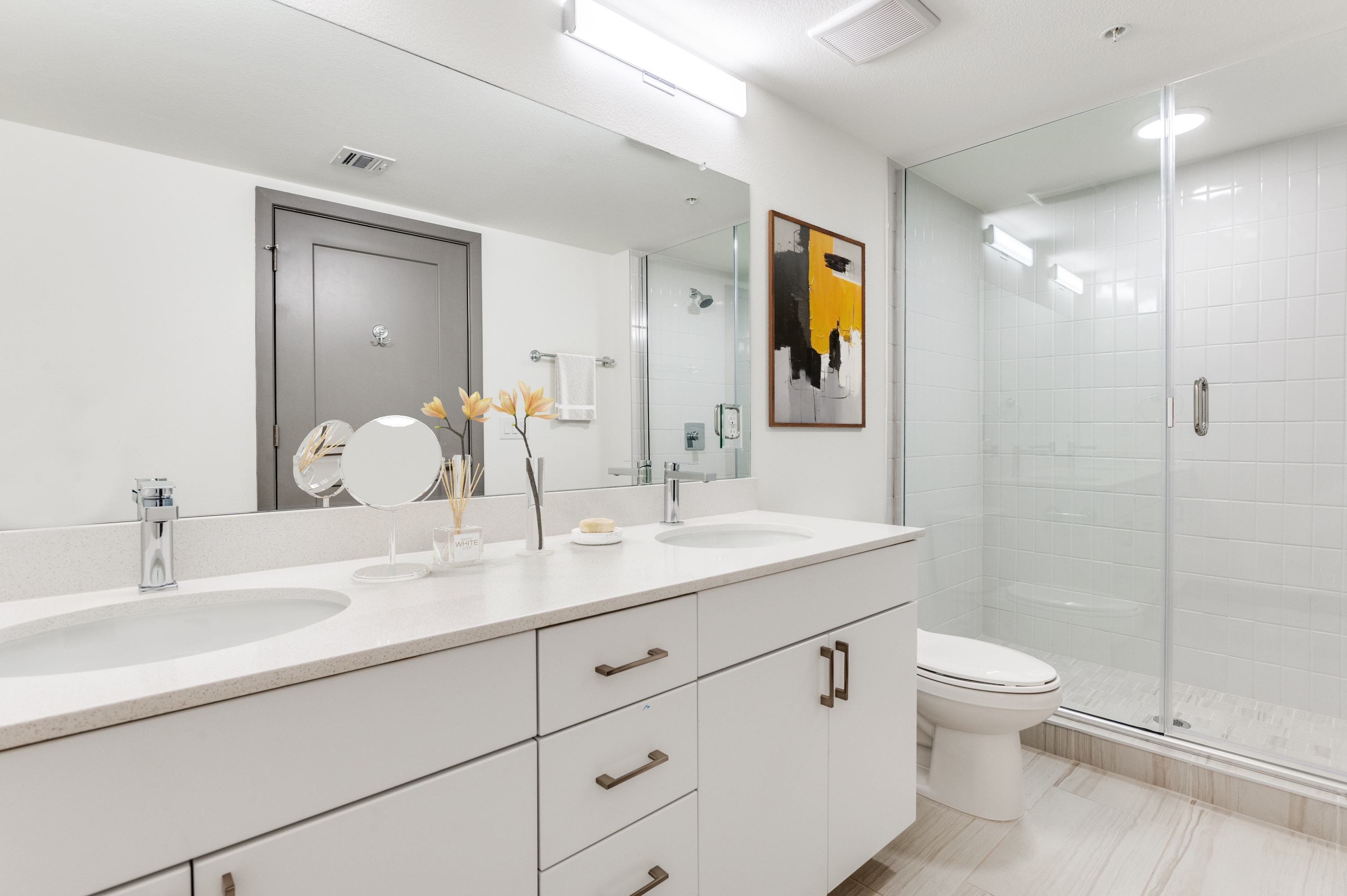 Modern bathroom with white cabinets, double sinks, a glass-enclosed shower, a toilet, wall mirror, and a yellow abstract painting—offering chic comfort in this Dallas Rental near Cabana Design District.