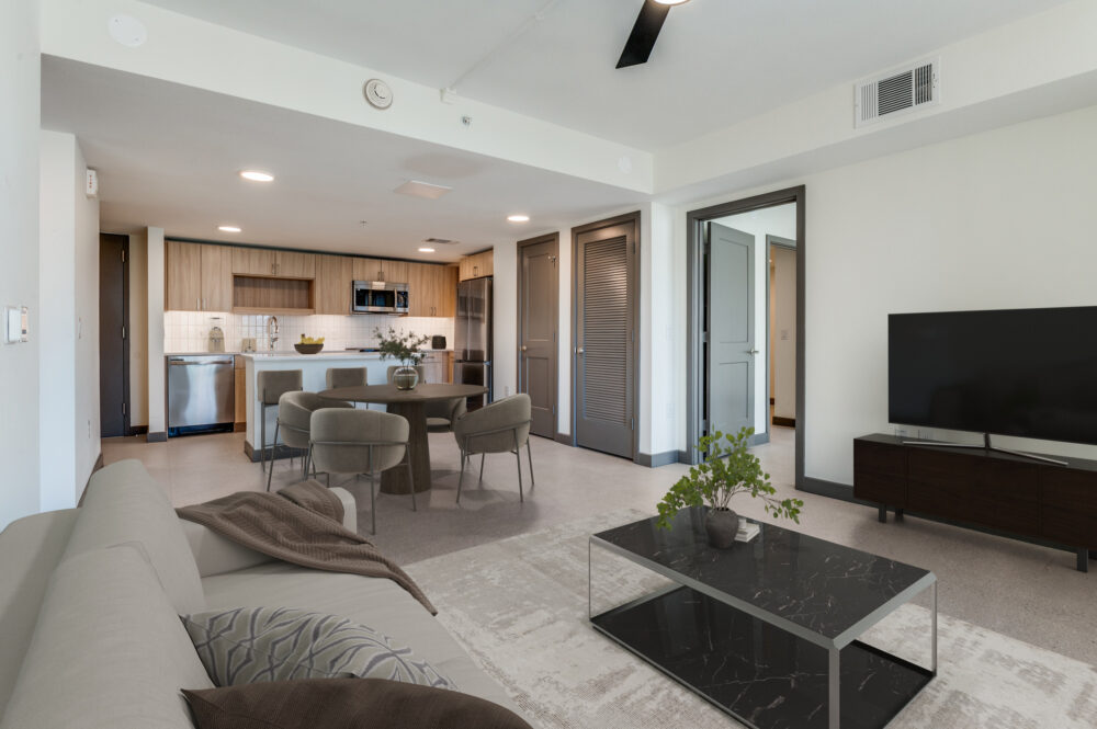 Modern open-plan living room and kitchen with neutral decor in a stylish Dallas rental, featuring a sofa, dining table, TV, and coffee table; doors lead to other rooms.