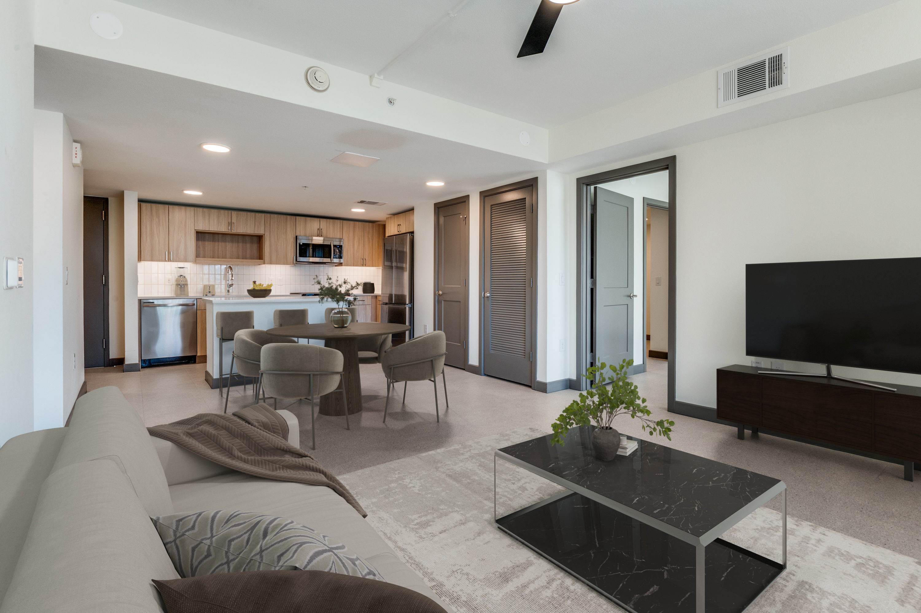 Modern open-plan living room and kitchen with neutral decor in a stylish Dallas rental, featuring a sofa, dining table, TV, and coffee table; doors lead to other rooms.