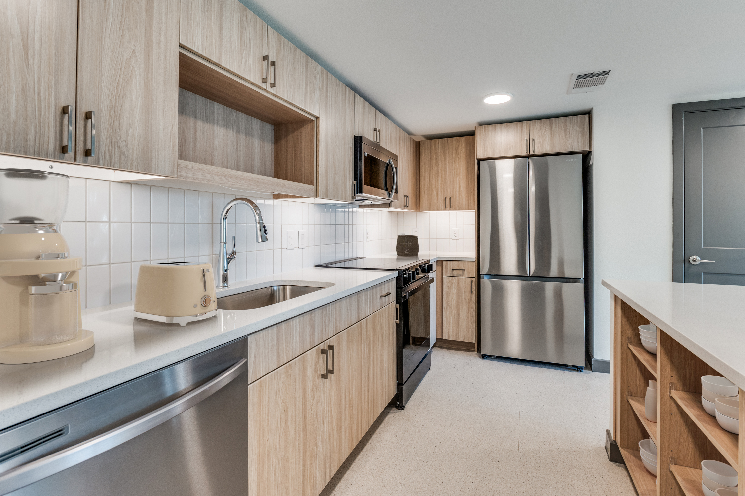 Modern kitchen in Dallas apartments with light wood cabinets, stainless steel appliances, white countertop, tile backsplash, and open shelving displaying dishes.