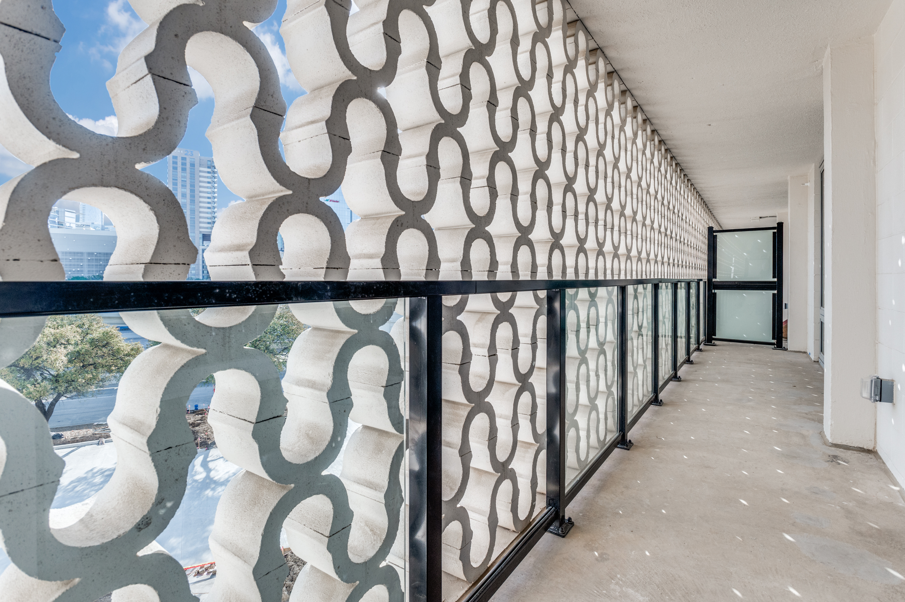 Outdoor apartment corridor with a decorative concrete privacy screen and glass railing, overlooking trees and distant buildings under a blue sky—perfect for enjoying views of historic Dallas apartments.