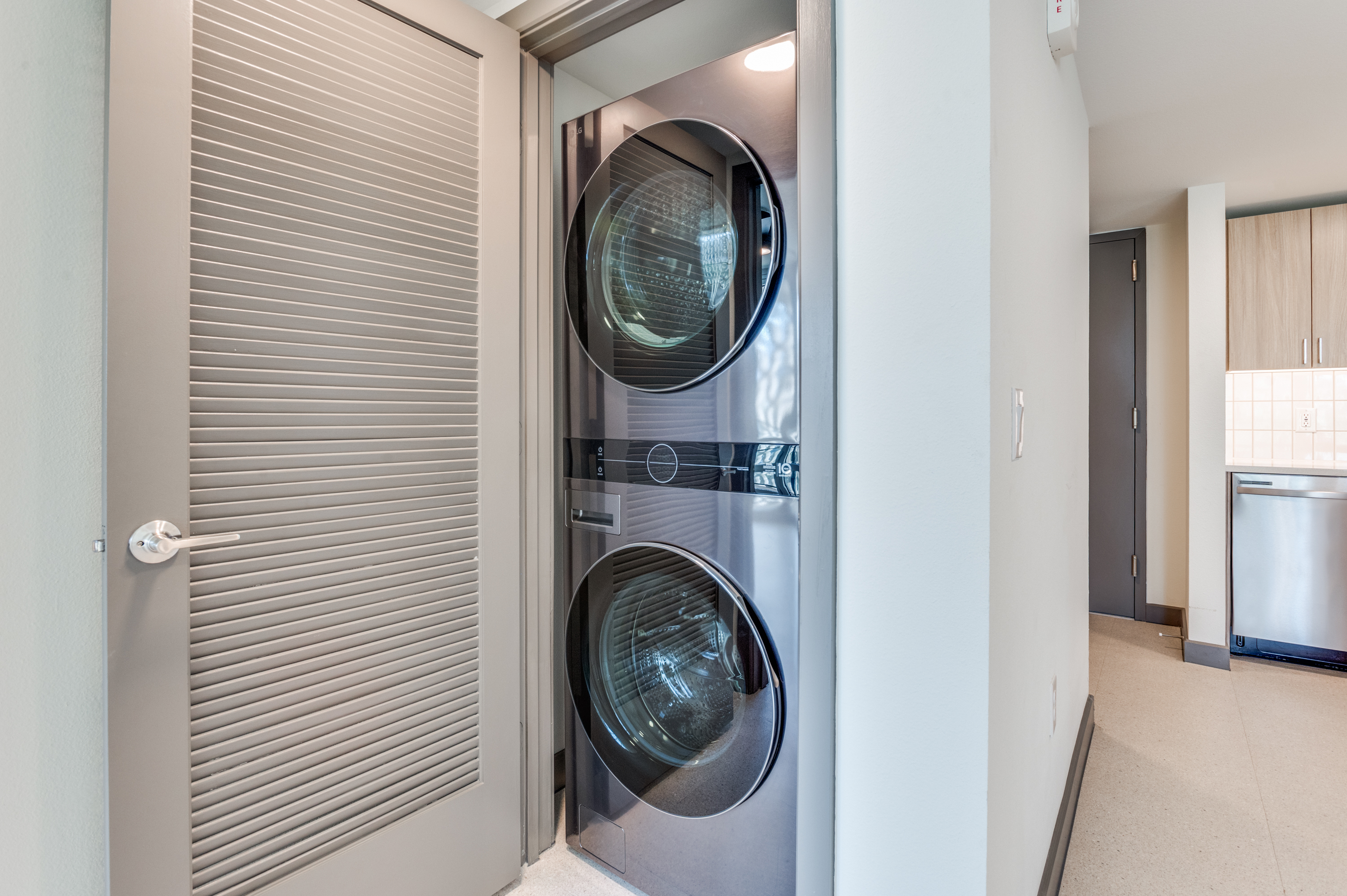 A stacked washer and dryer unit is installed in a closet with louvered doors, adding convenience to the hallway of these modern Dallas Apartments.