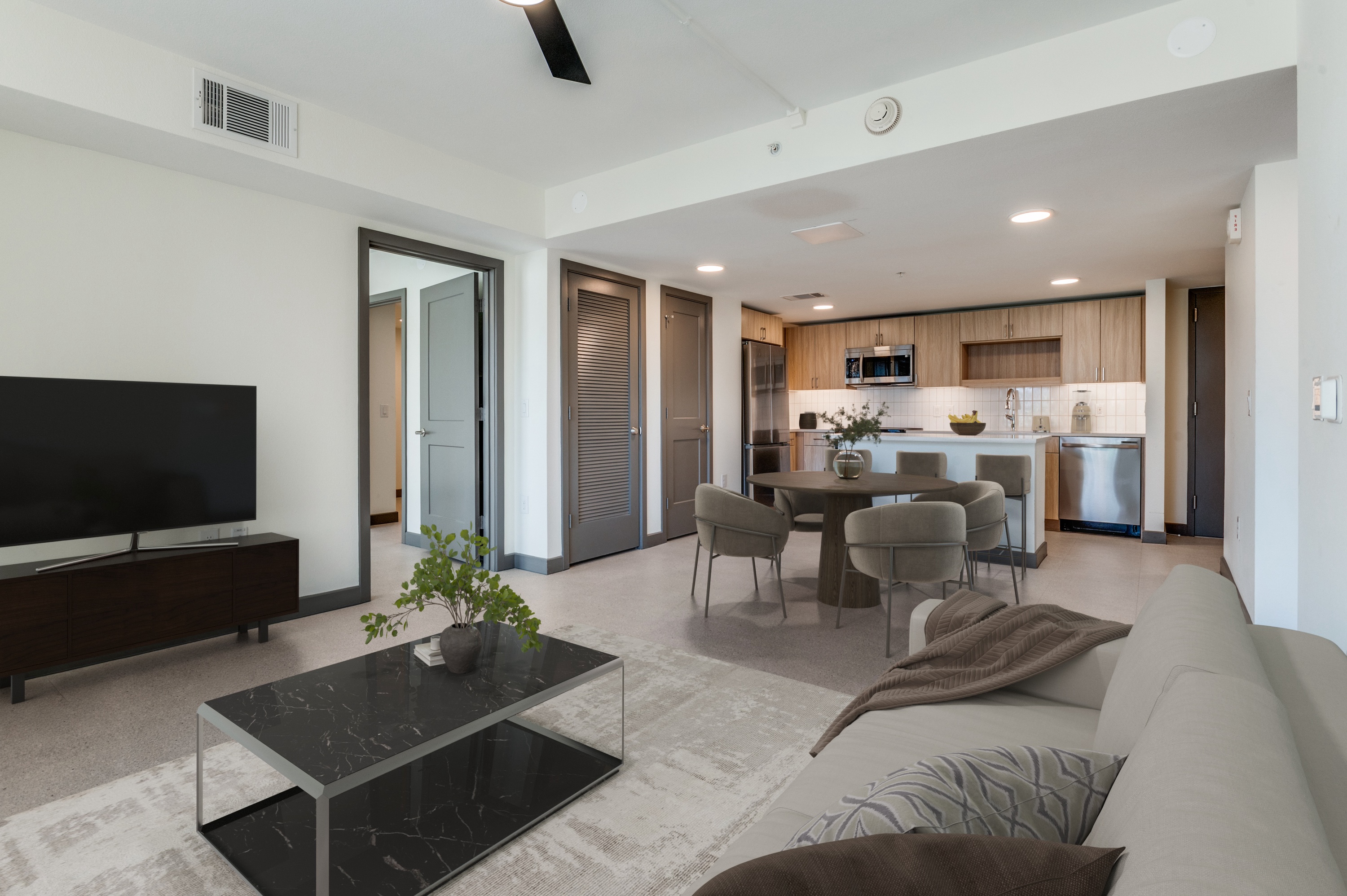 Modern Dallas Rental with a spacious living room and kitchen area featuring a sofa, coffee table, TV, dining table, and stainless steel appliances. Neutral tones and minimalistic decor define this Cabana Design District home.