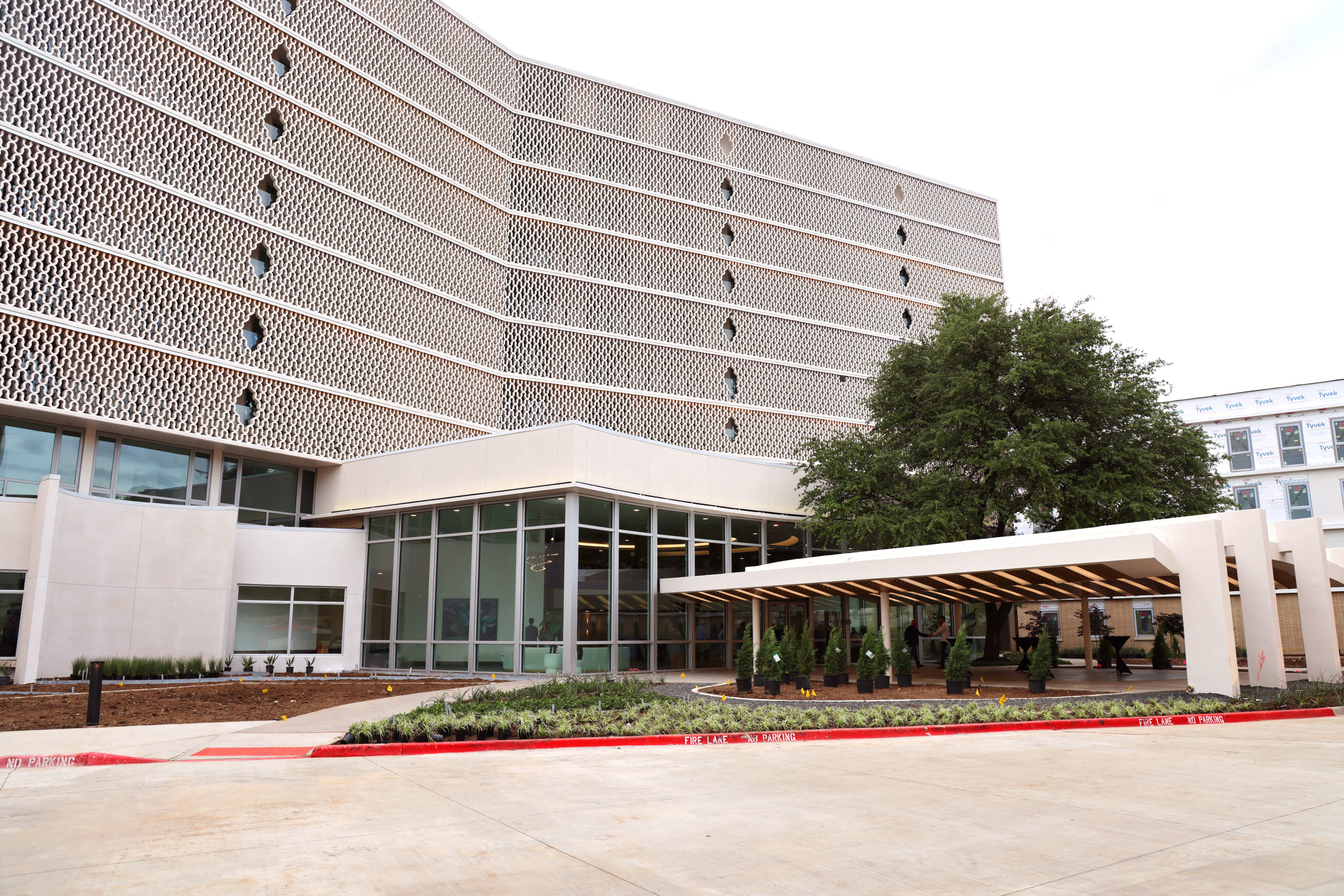 A multi-story modern building with geometric patterned façades, large windows, landscaped greenery, and a covered walkway near the entrance—perfect for those seeking stylish Dallas Rental apartments.