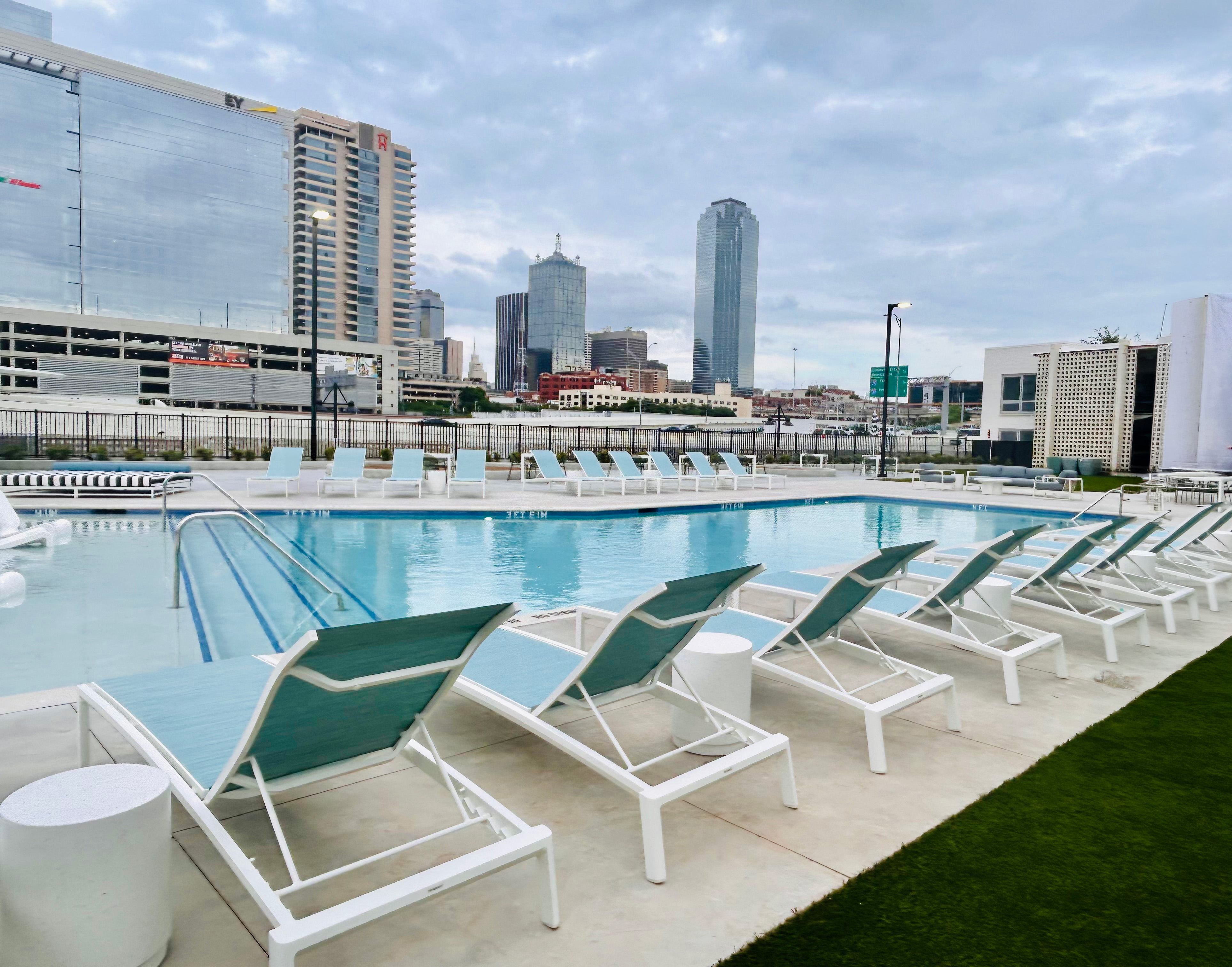 A rooftop swimming pool with lounge chairs overlooks the historic Dallas skyline, offering stunning views under a cloudy sky—perfect for relaxation in the heart of the Cabana Design District.