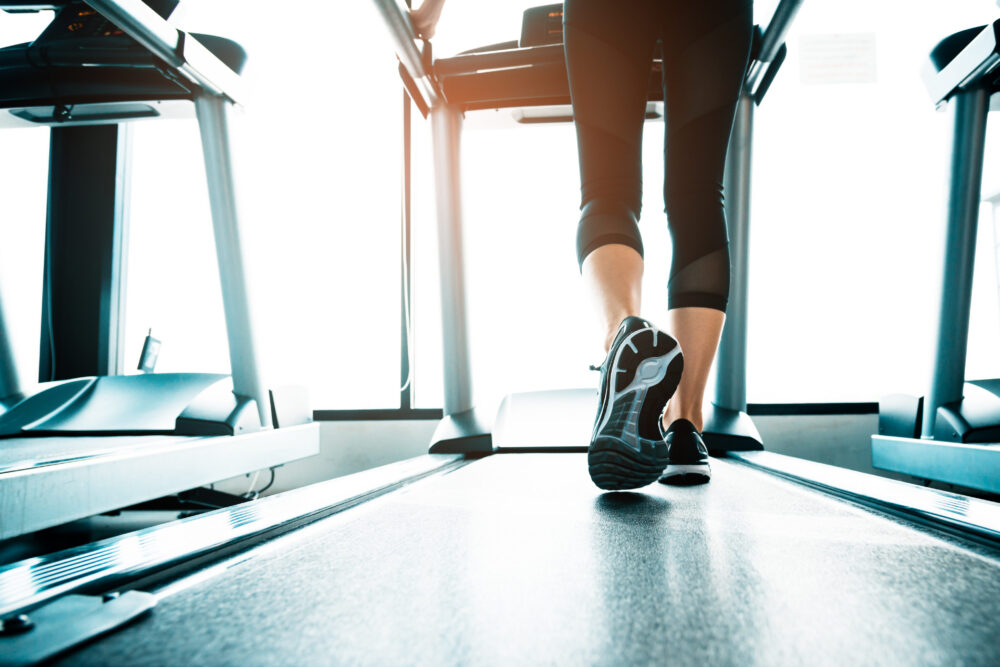 Person walking on a treadmill in a gym in Dallas, shown from behind at foot level, wearing athletic shoes and black leggings. Other treadmills are visible beside them in this modern Cabana Design District setting.