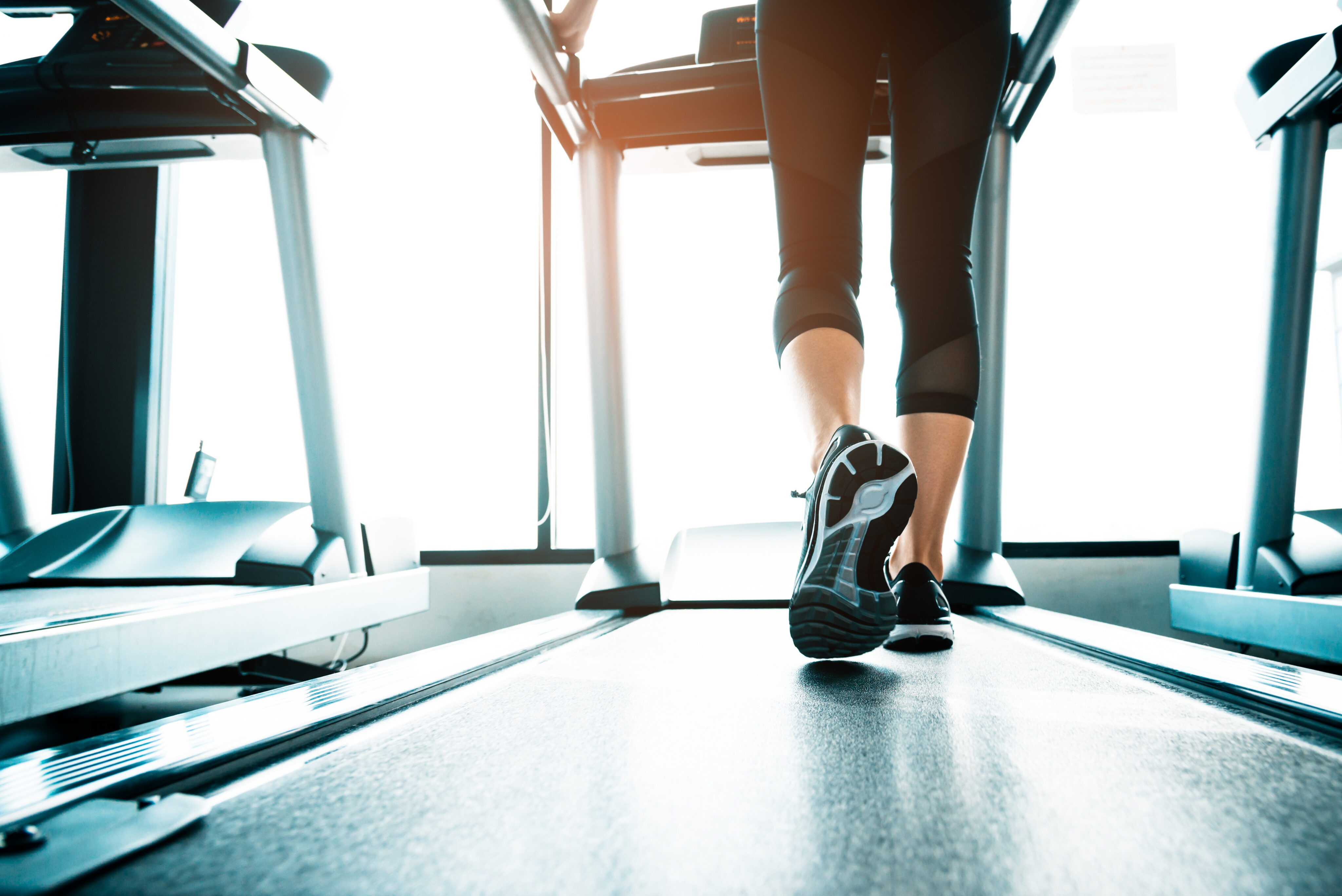 Person walking on a treadmill in a gym in Dallas, shown from behind at foot level, wearing athletic shoes and black leggings. Other treadmills are visible beside them in this modern Cabana Design District setting.